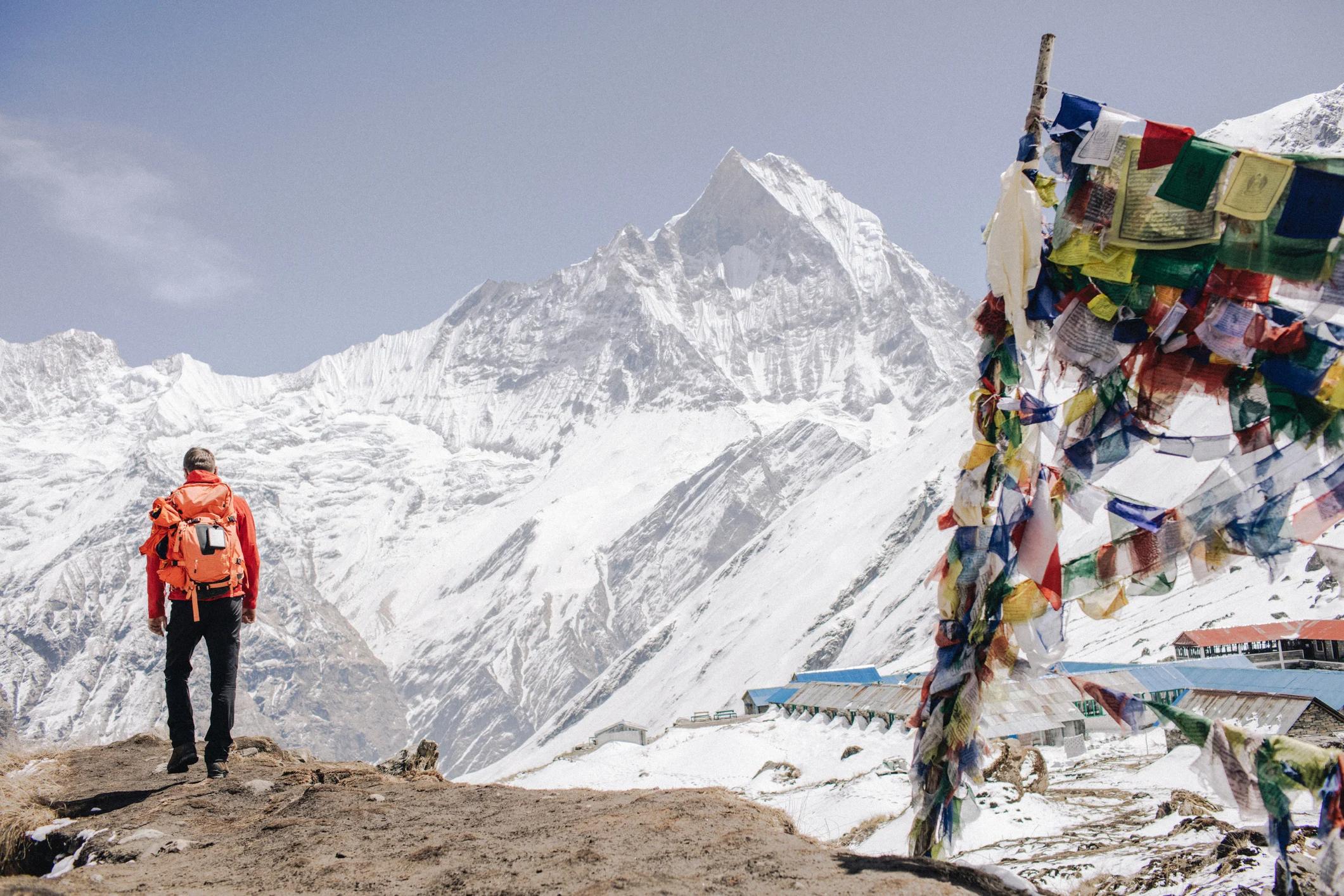 Hiker in an orange jacket stands on a rocky outcrop, surrounded by snow-capped mountains and colorful prayer flags.