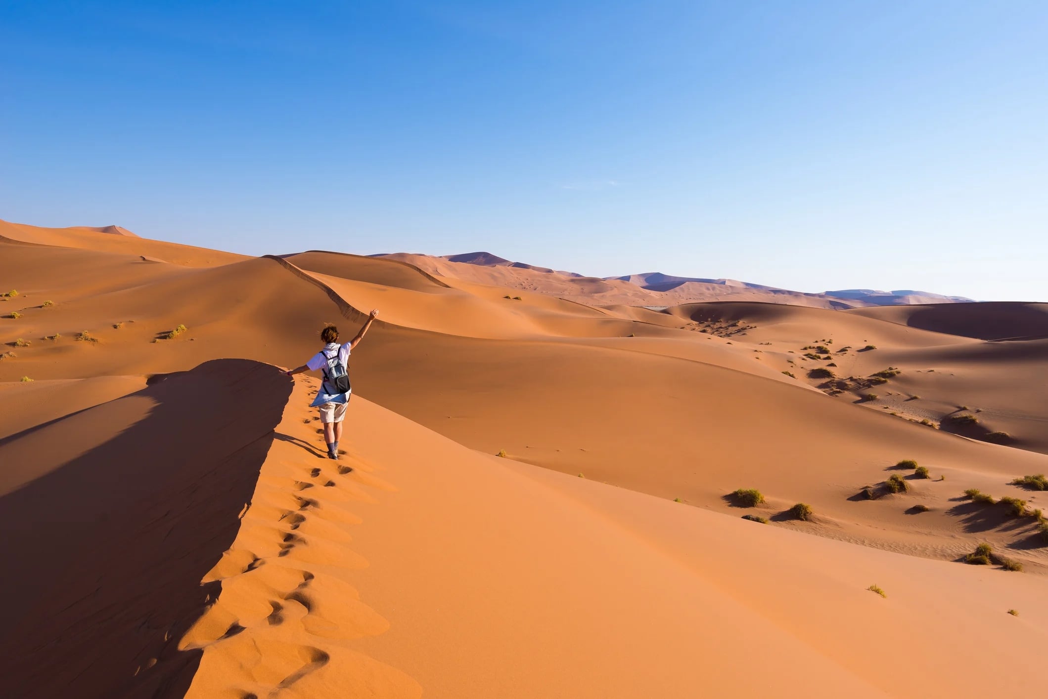 A person walks on a sandy dune in a desert, with rolling hills and a clear blue sky in the background.