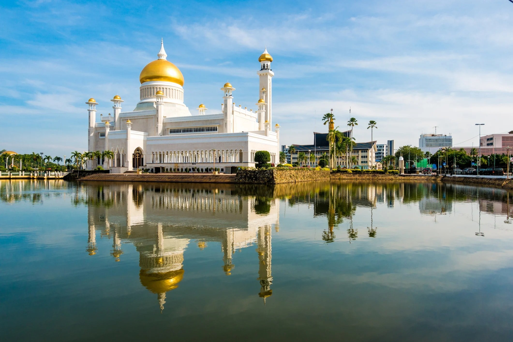 A grand white mosque with a gold dome reflects in a tranquil lake, under a blue sky with scattered clouds.