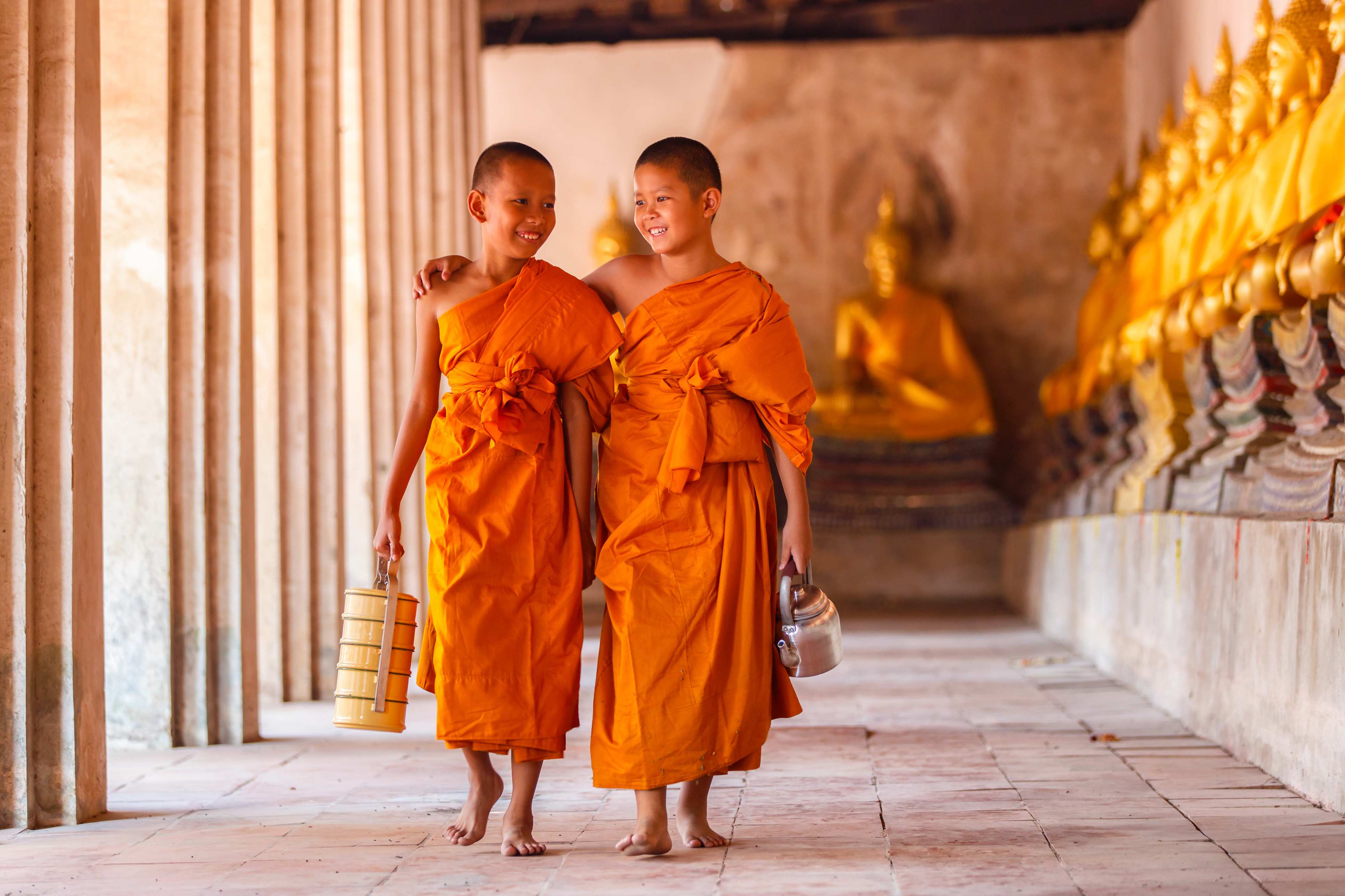 Two young monks in orange robes walking together, smiling, with golden statues in the background.