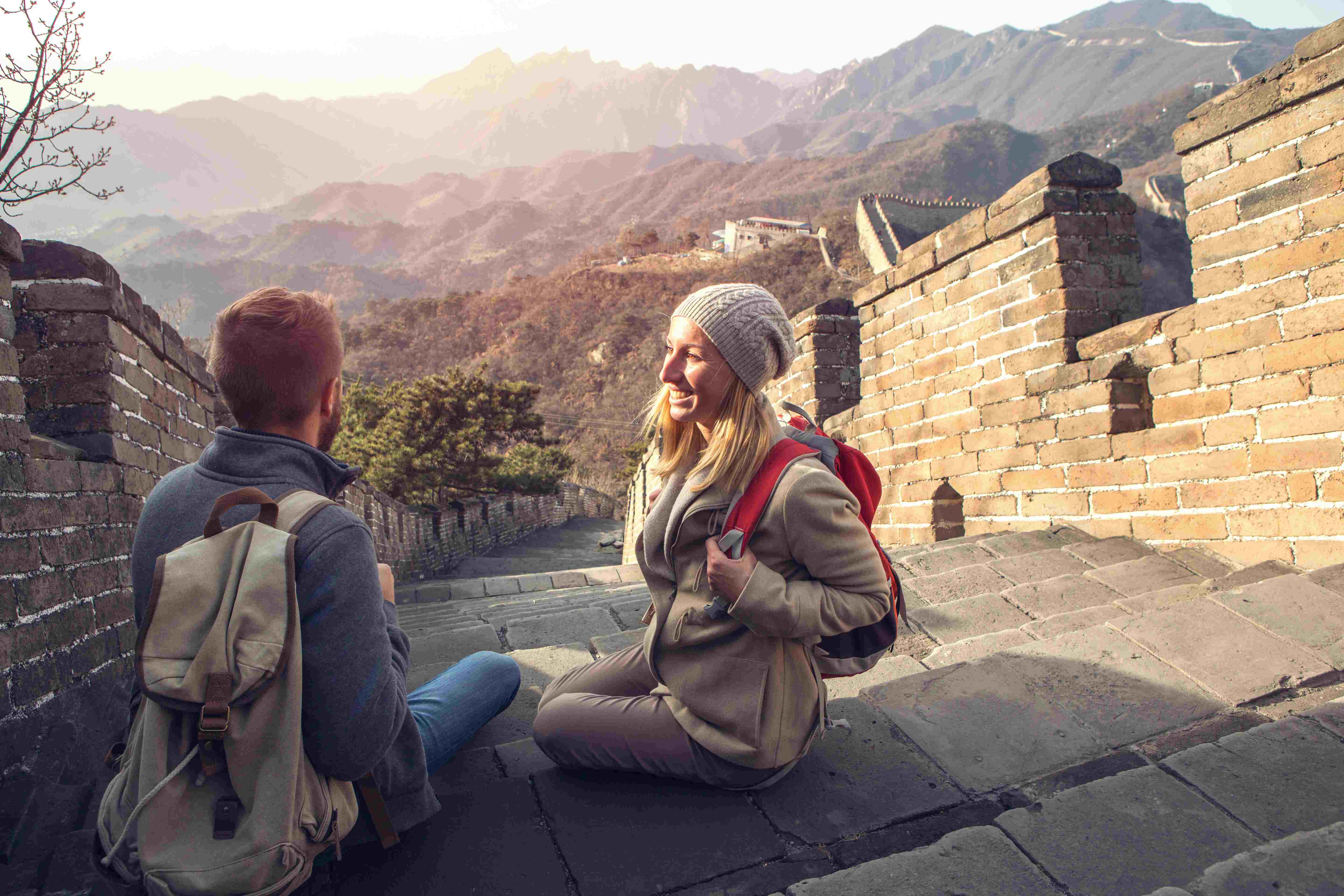 Two hikers sit on the Great Wall of China, enjoying the view of mountains in the background during sunset.