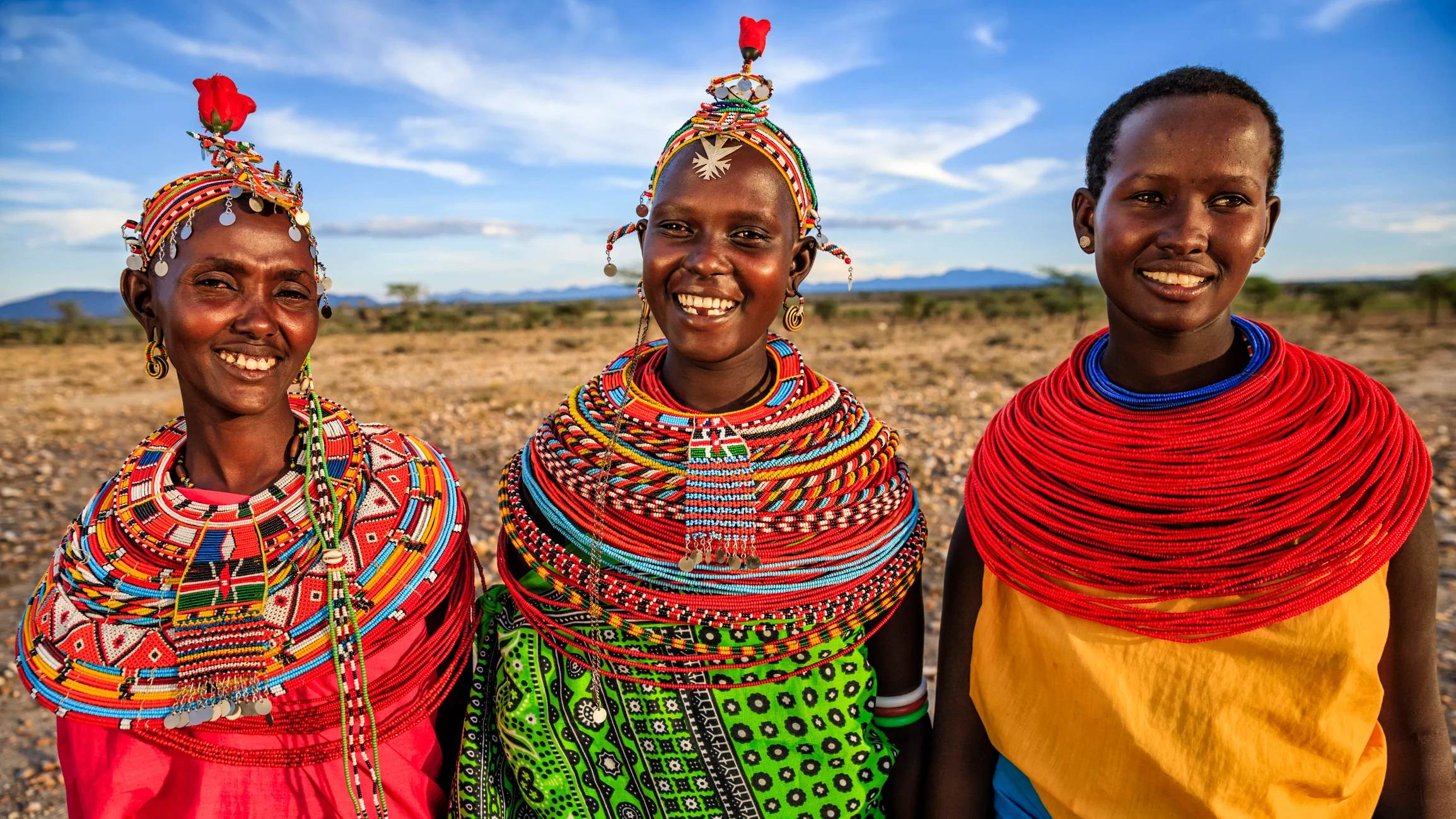 Three smiling women in colorful traditional attire stand together against a scenic landscape.