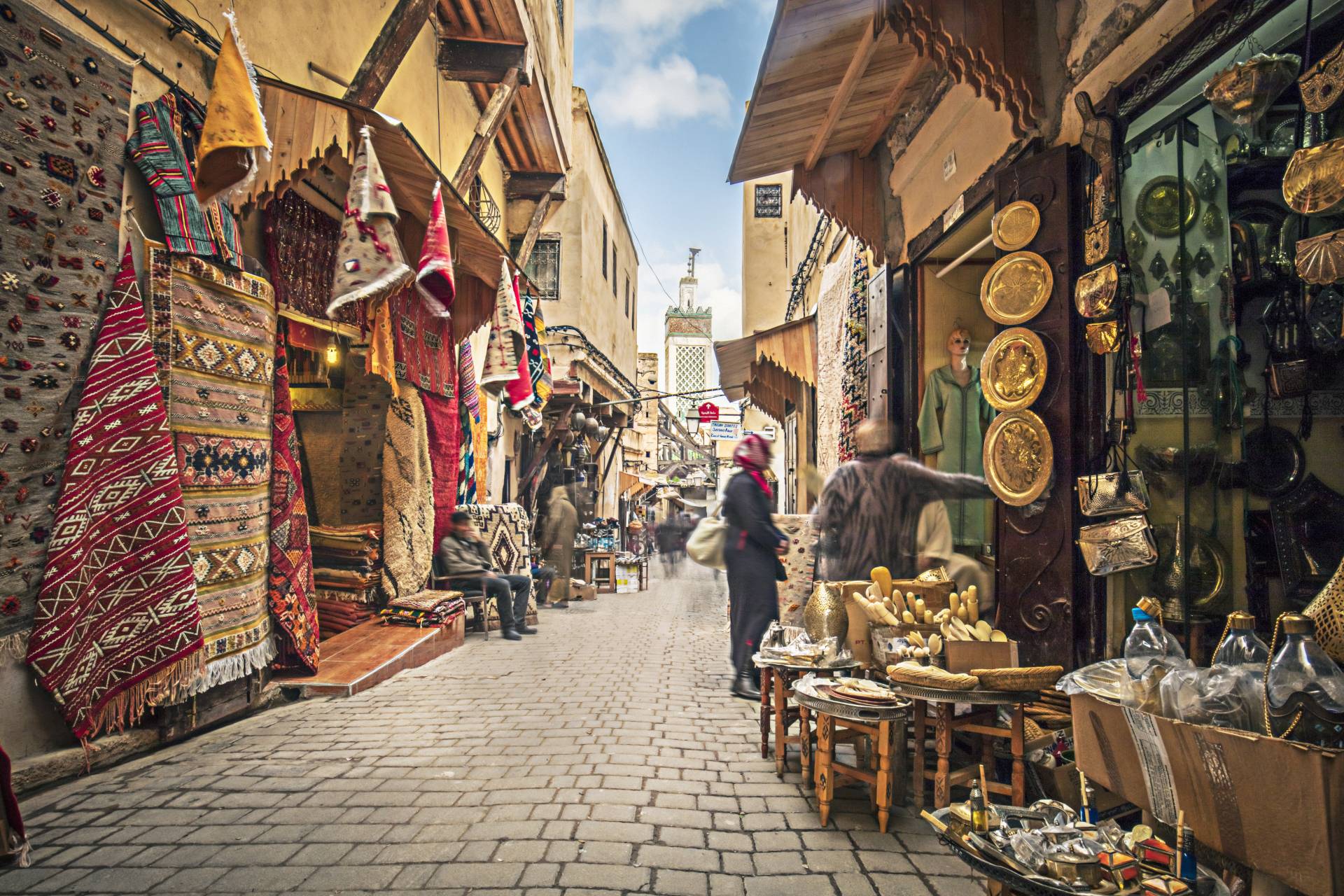 Narrow market street lined with colorful rugs, shops, and people browsing various goods under a clear blue sky.