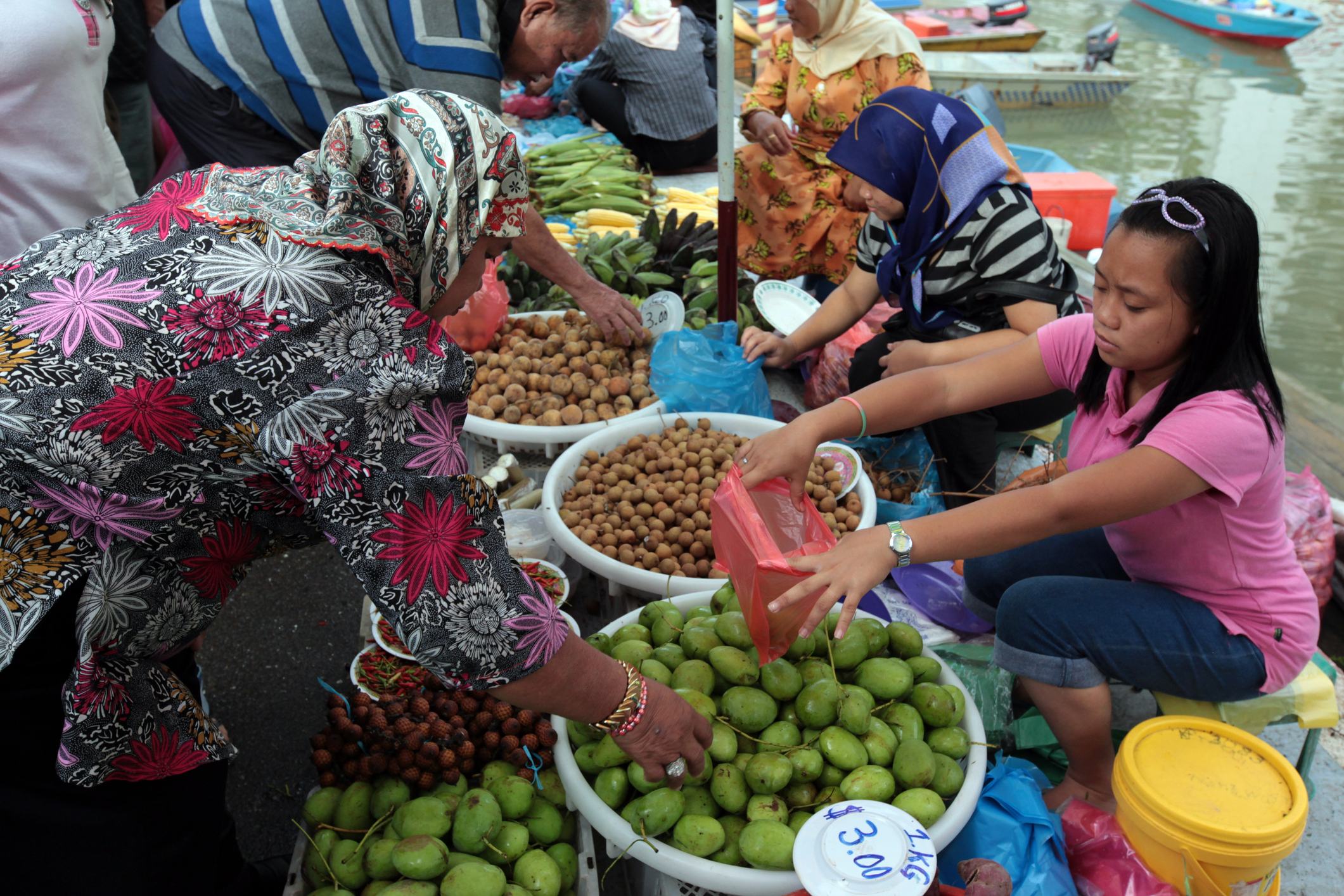 A vibrant market scene with women selling fruits from baskets along a waterway, showcasing various colorful produce.