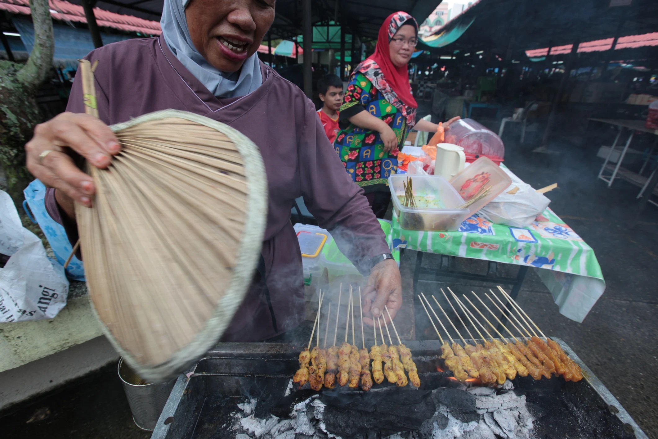 A vendor fans smoky grilled skewers at a market, with people and colorful food stalls in the background.