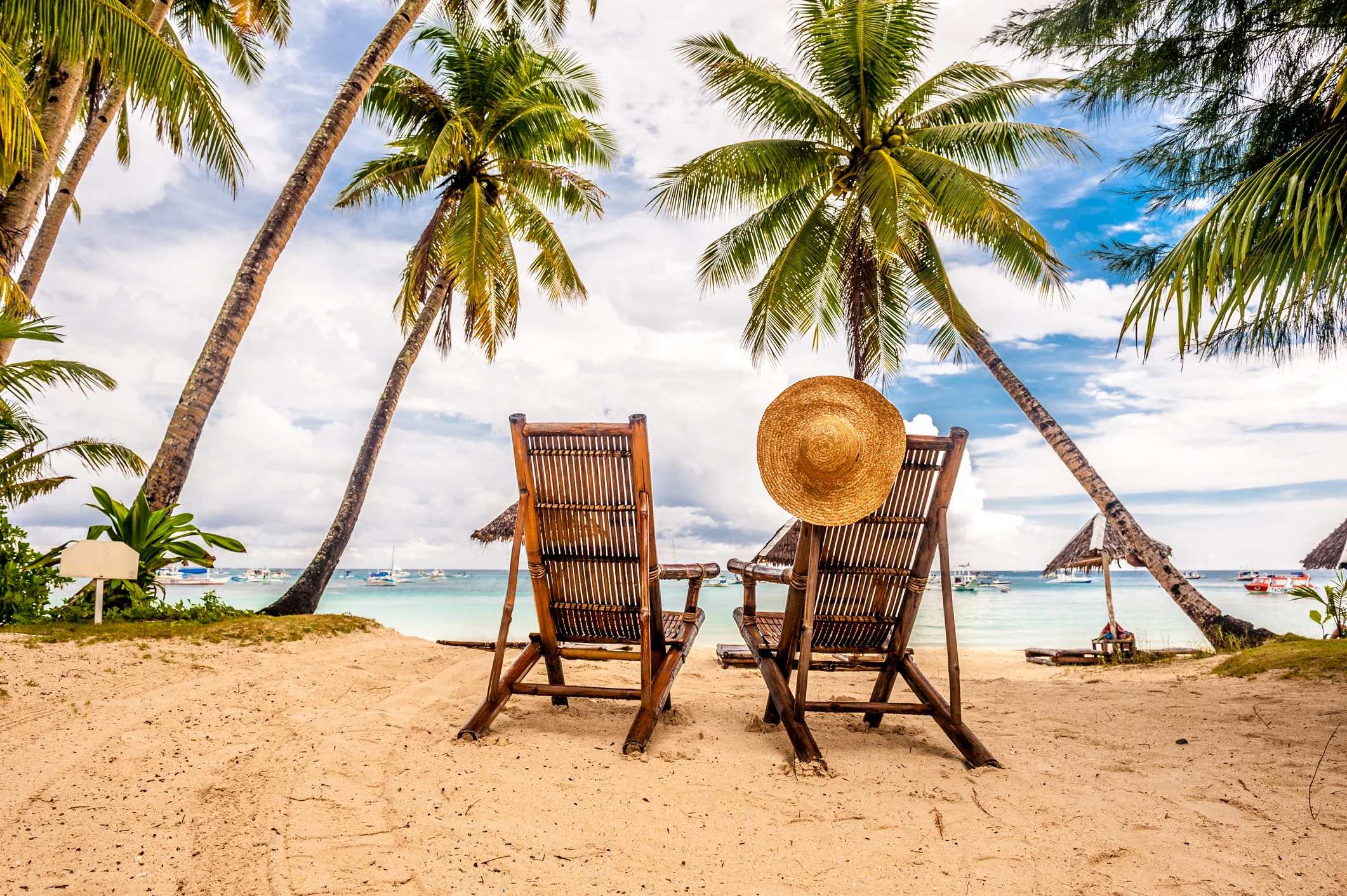 Two wooden lounge chairs on a sandy beach, framed by palm trees, with a straw hat resting on the back of one chair.