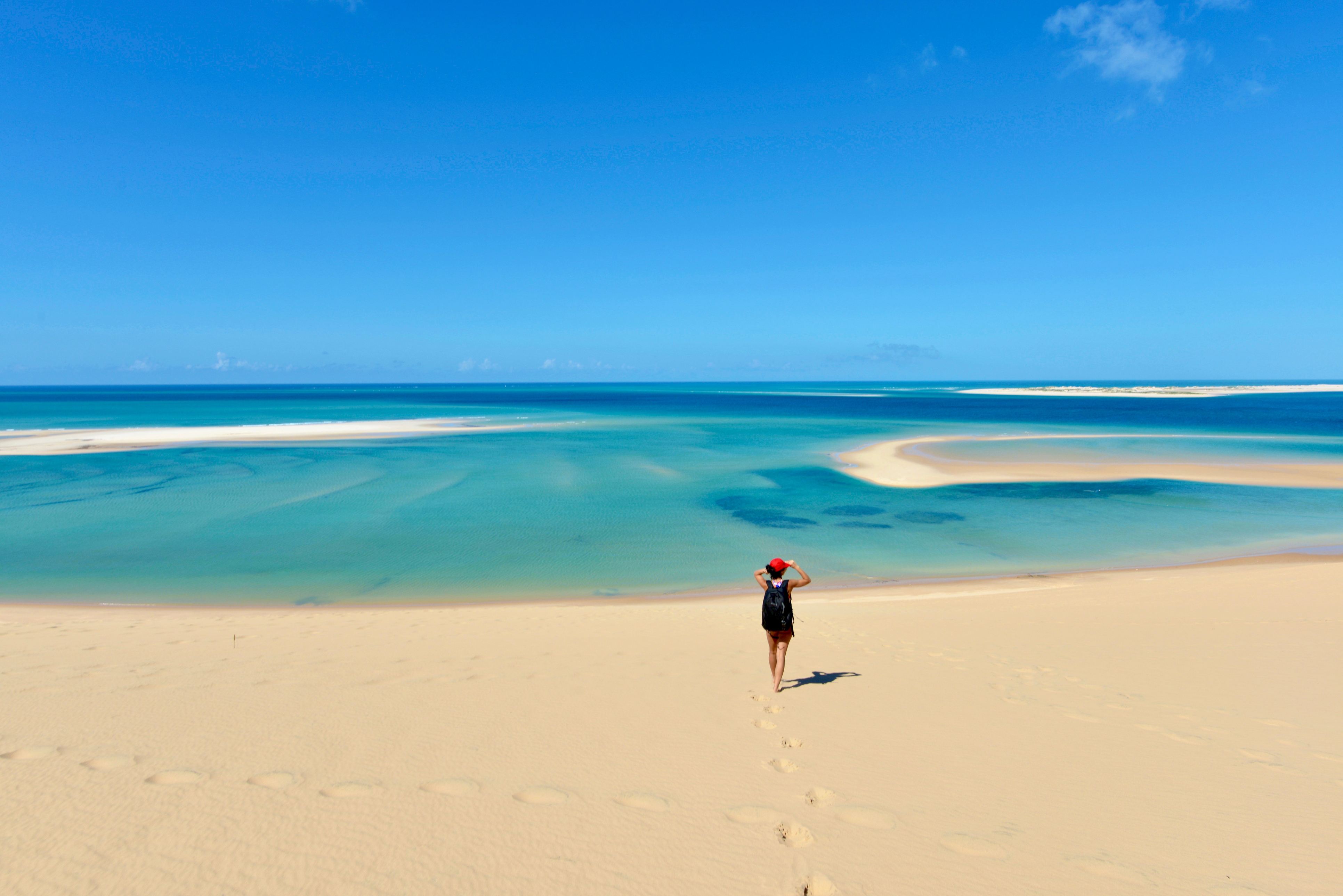A person walks along a sandy beach, with clear blue water and a bright sky in the background.