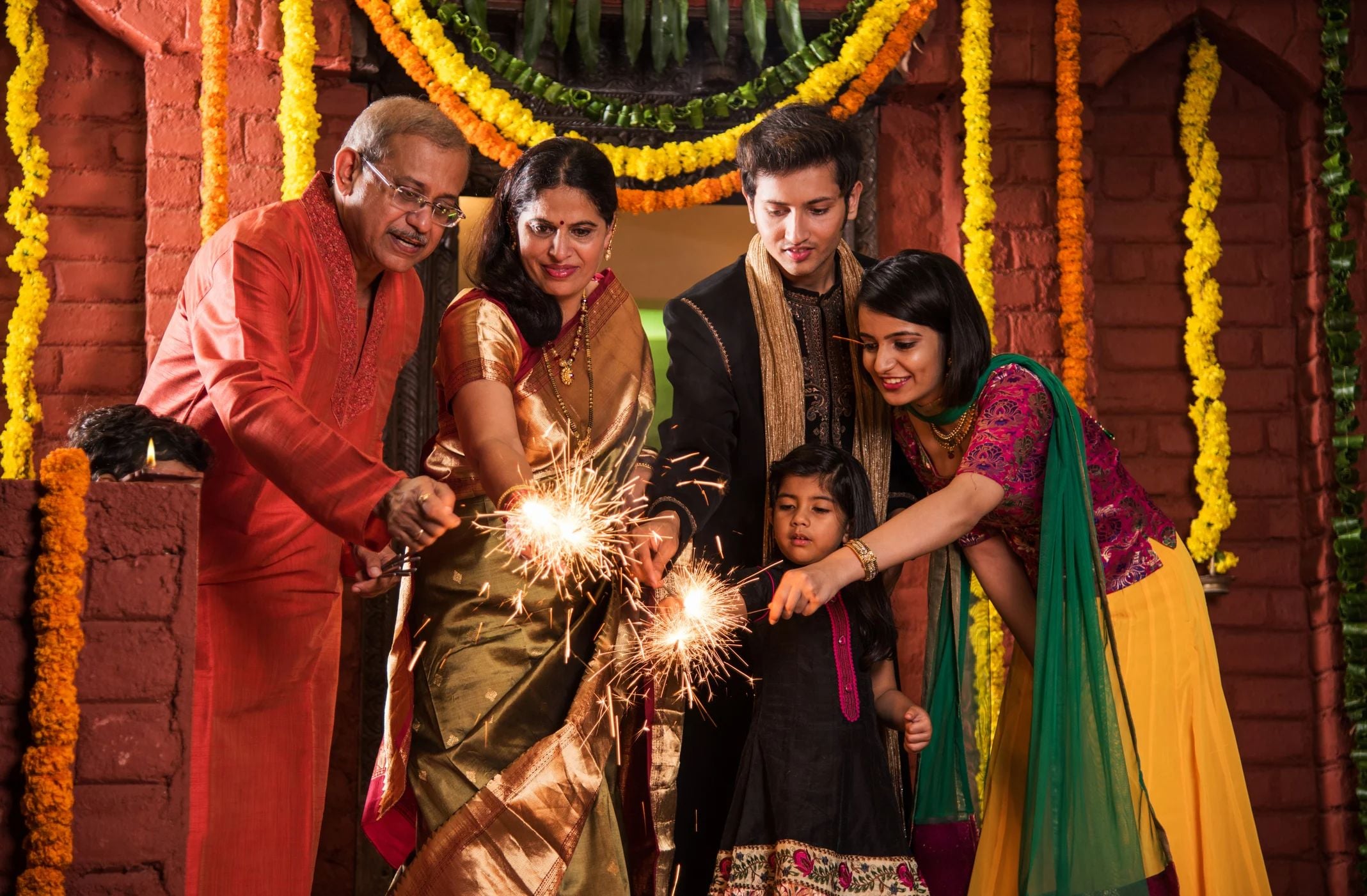 A family celebrates together, lighting sparklers in traditional attire surrounded by colorful flowers and festive decorations.