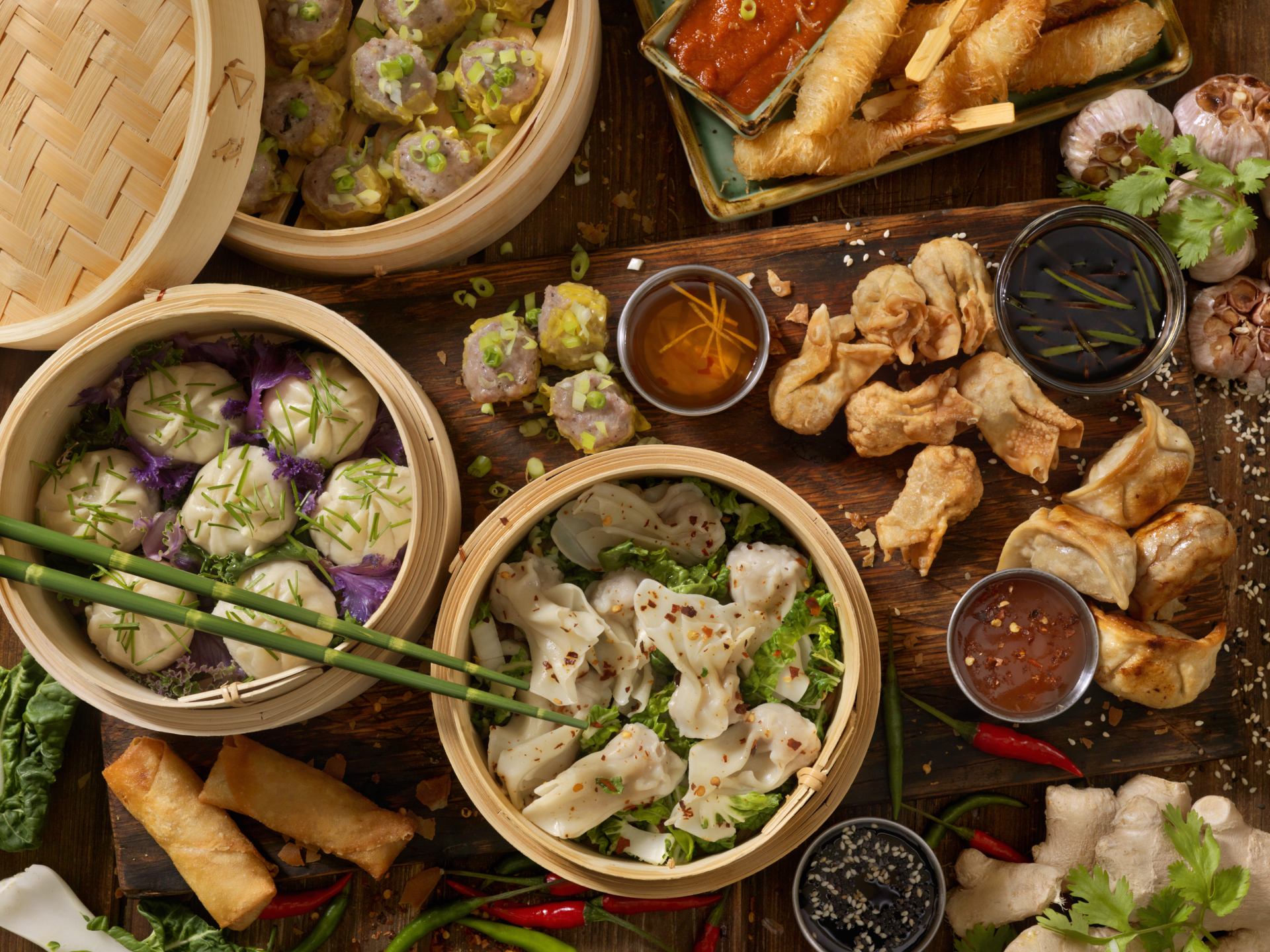 A spread of various Asian dumplings and spring rolls, served in bamboo steamers and accompanied by dipping sauces.