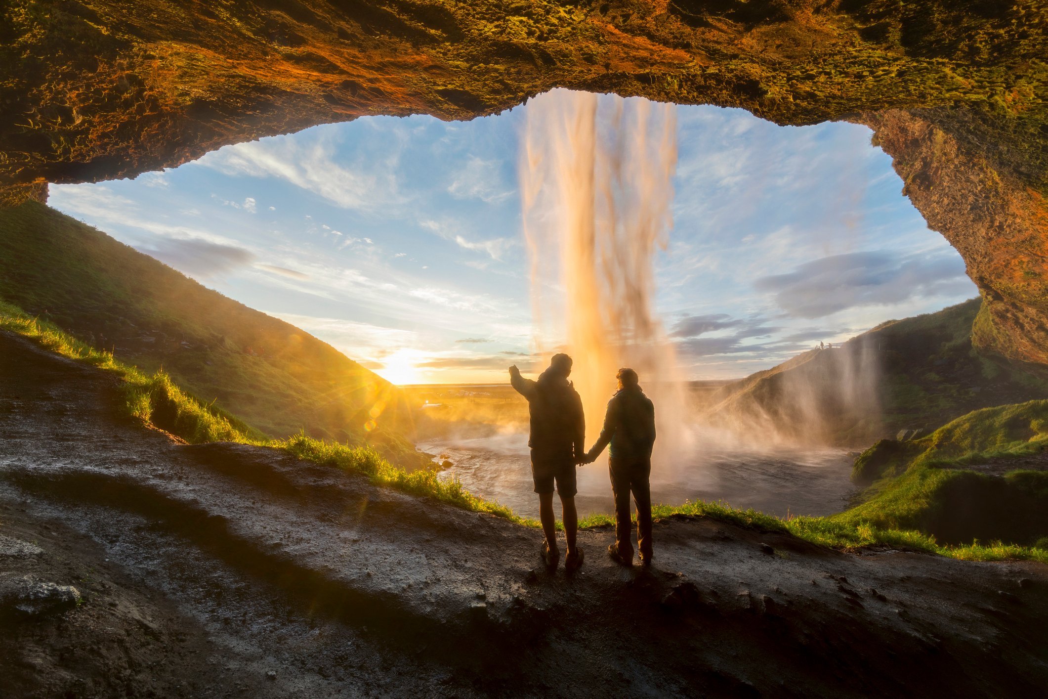 Discover the magic of Seljalandsfoss, Iceland, with those who matter most.  Tourists under waterfall in Seljalandsfoss, Iceland, man pointing with finger to the sun