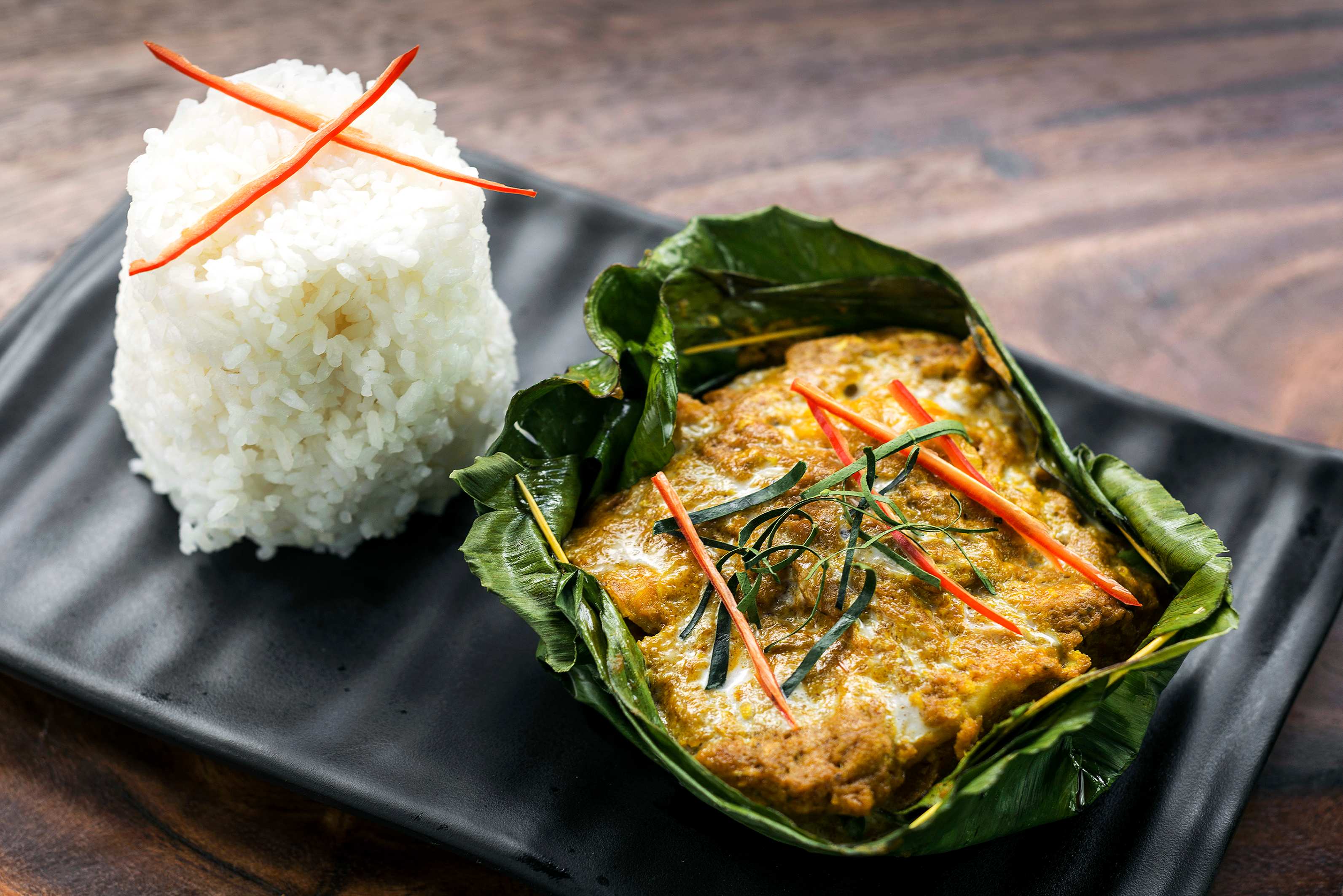 A dish featuring fish curry in banana leaves, garnished with herbs and red chilies, served with a side of white rice.