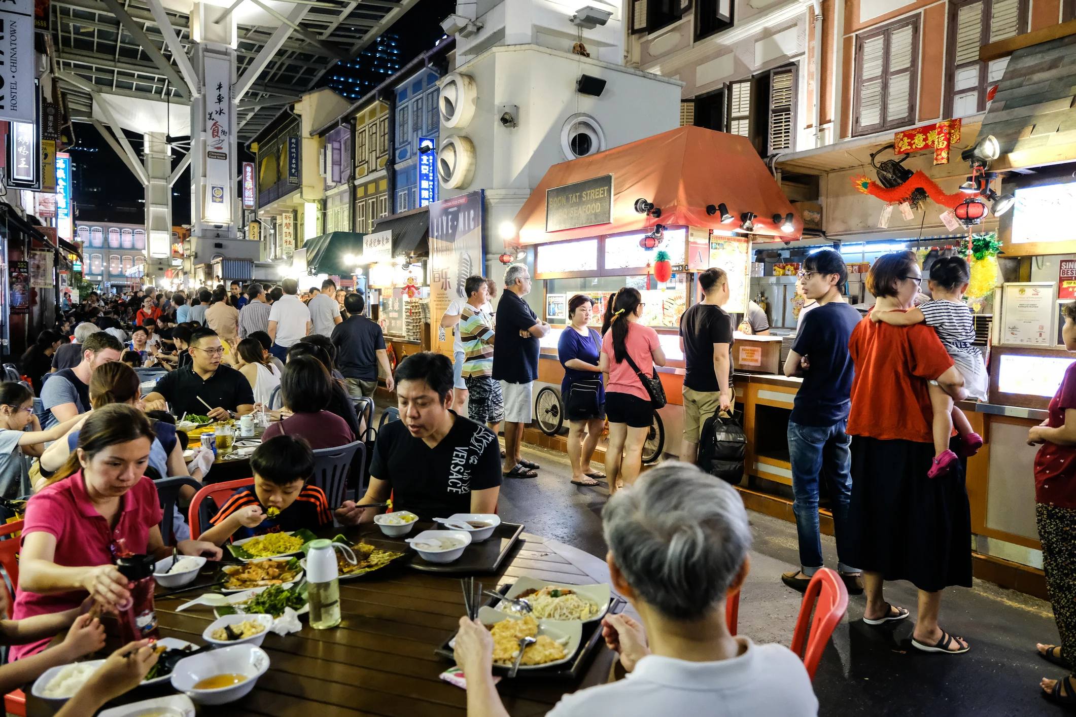 Busy night market scene with people dining and standing near food stalls, surrounded by colorful shopfronts.