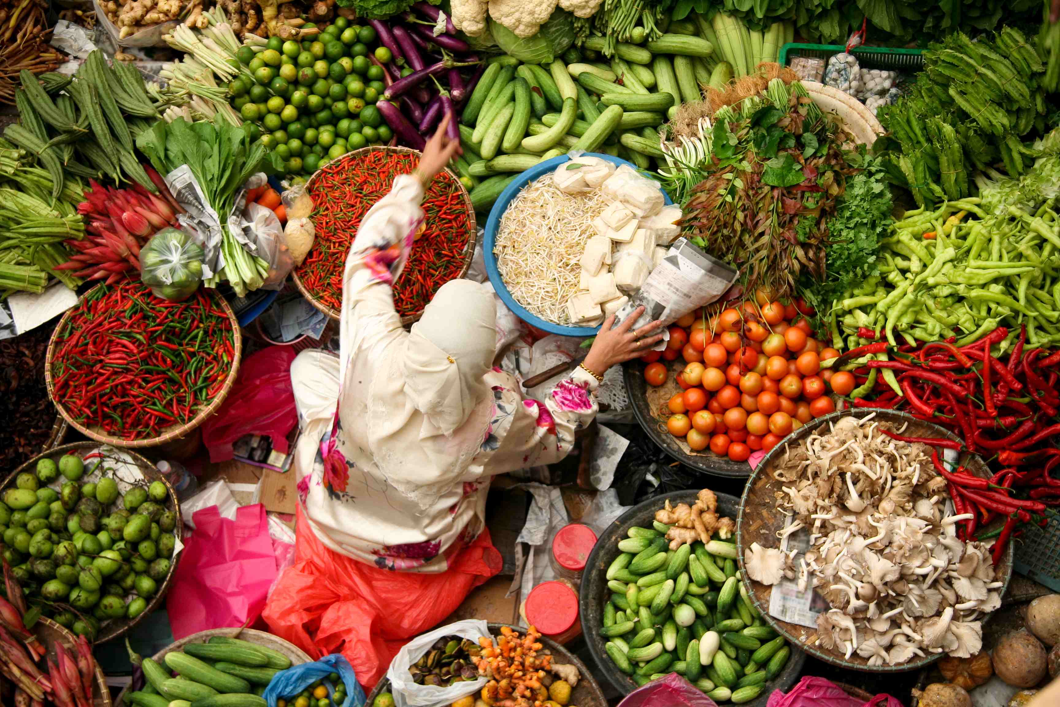 A vendor in a market reaches for produce among vibrant displays of fruits and vegetables in various baskets.