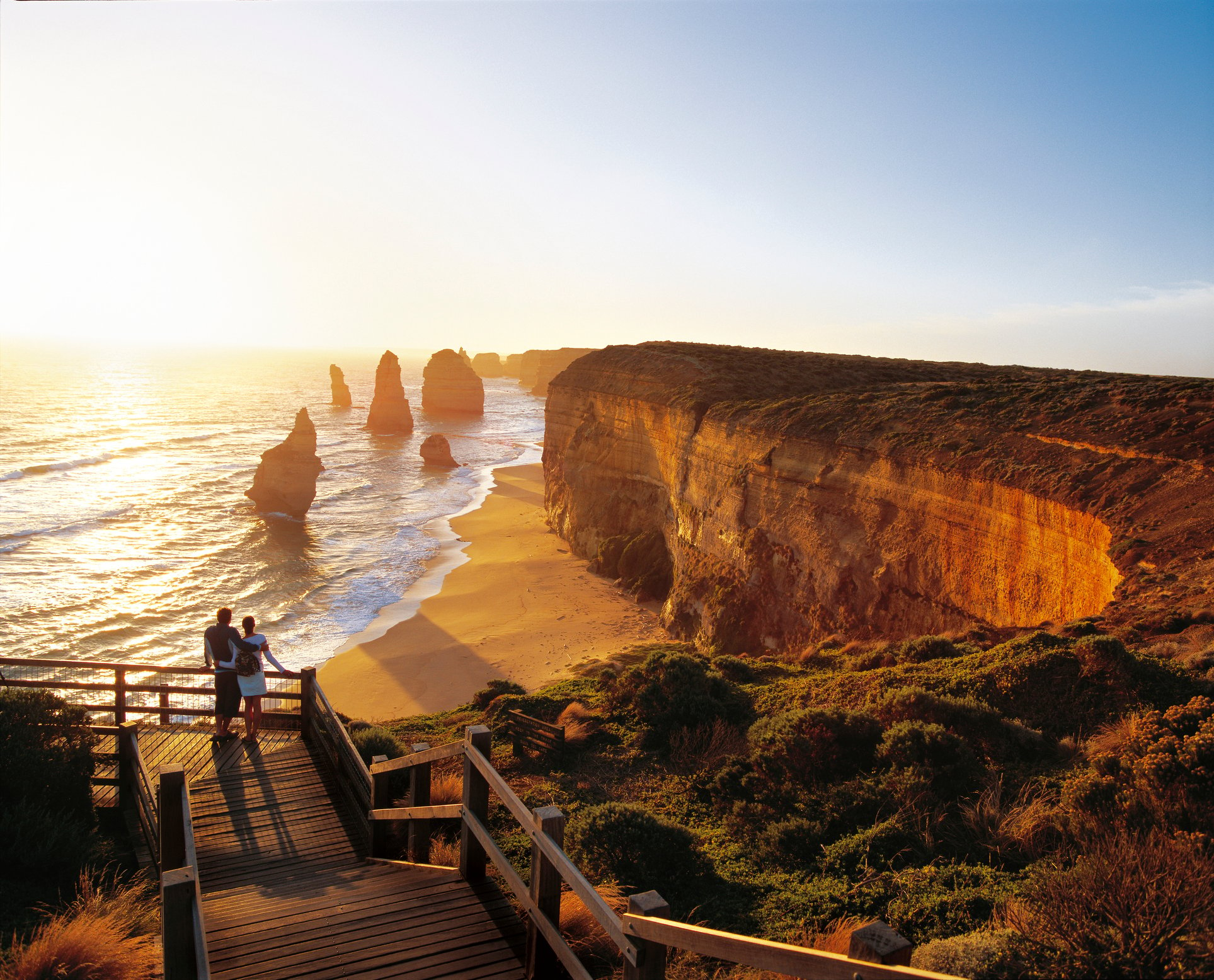 A couple stands on a wooden walkway overlooking a beach with cliffs and sea stacks at sunset.