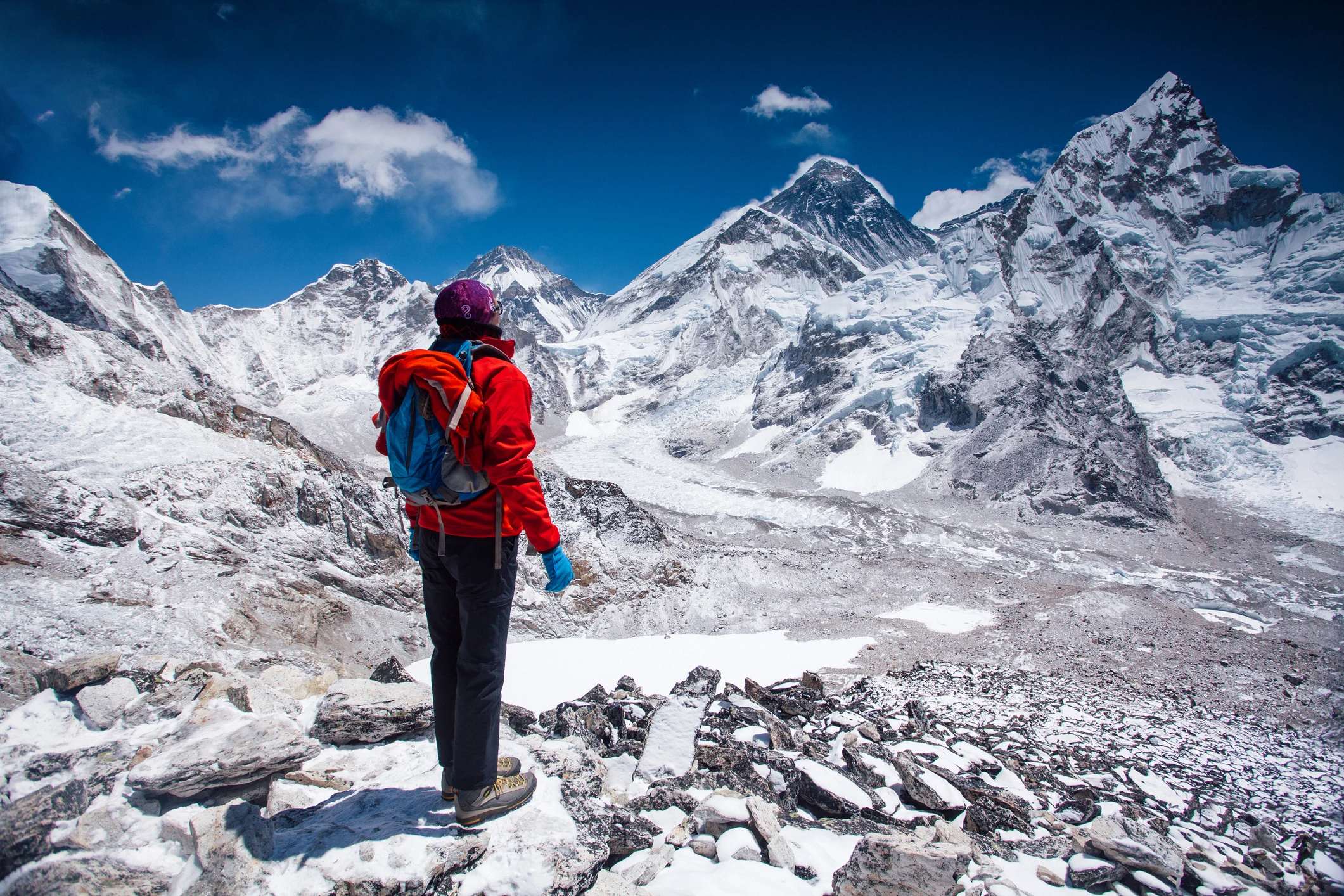 Person in a red jacket stands on rocky terrain, overlooking snow-capped mountains under a clear blue sky.