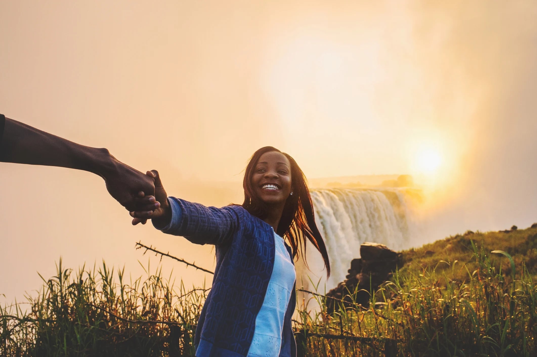 A woman joyfully holds hands with a person, standing near a waterfall at sunset amidst lush greenery.