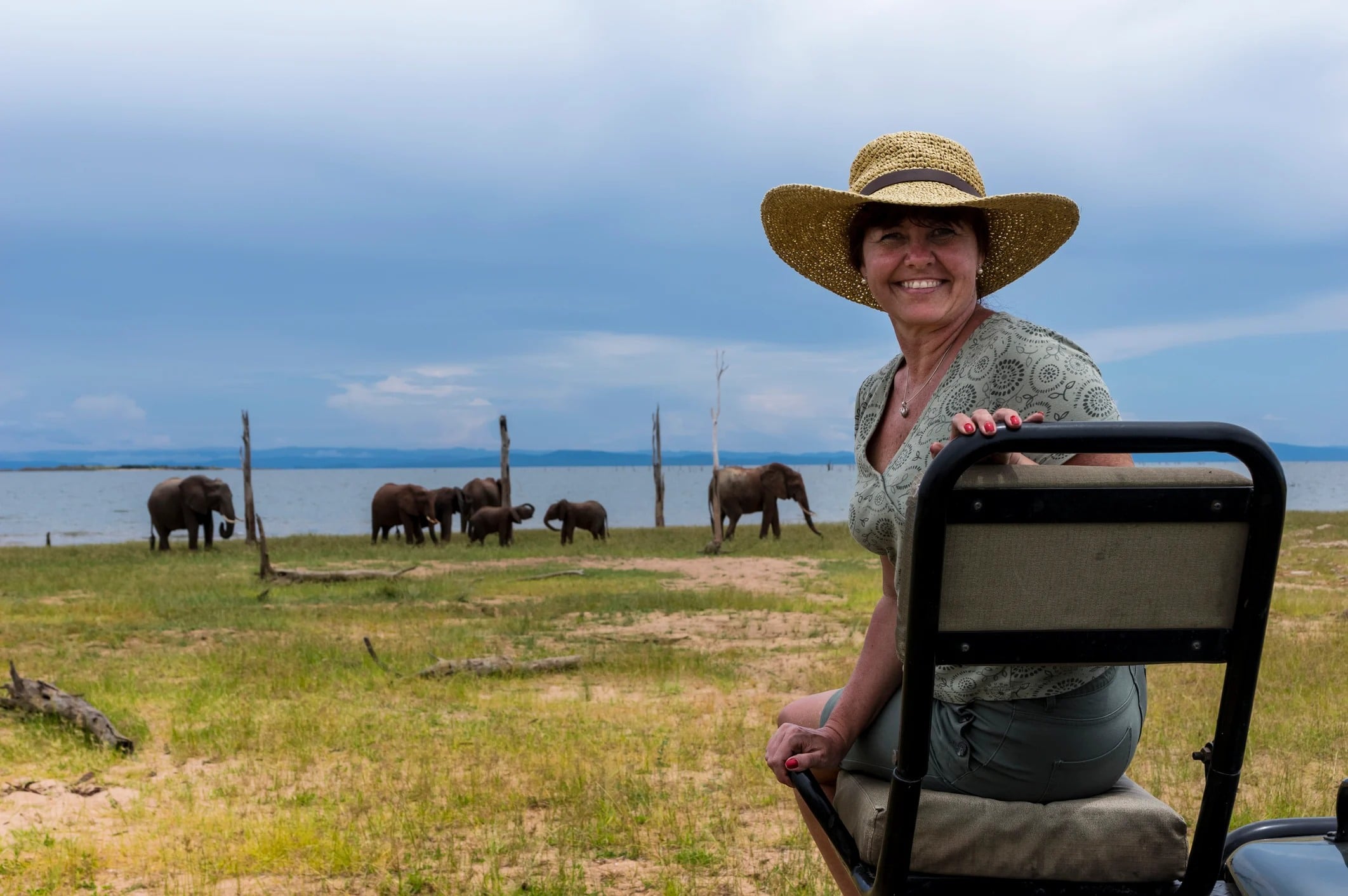 A woman in a straw hat smiles while seated, with elephants grazing in a landscape by a lake under a cloudy sky.