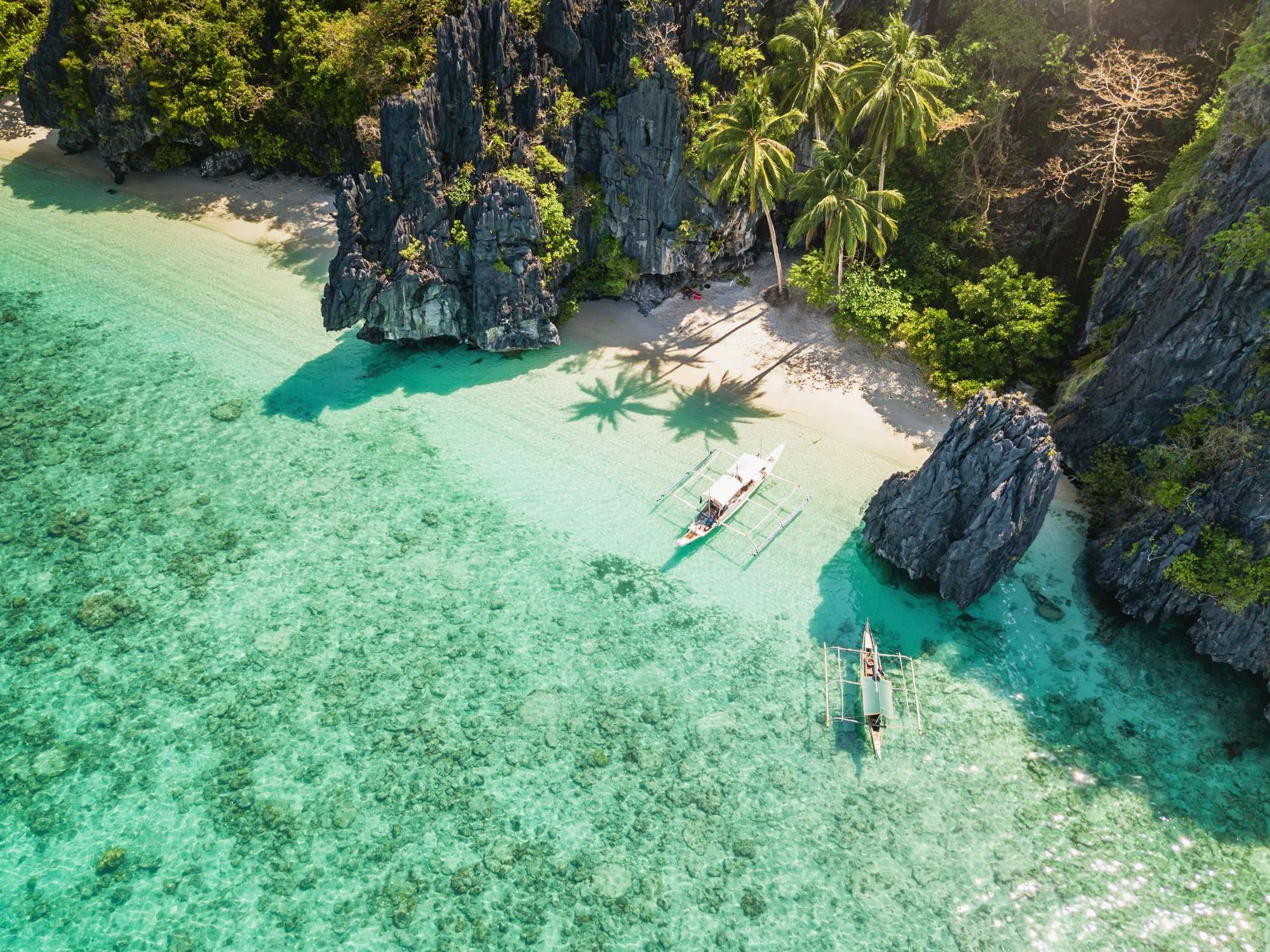 Aerial view of a clear turquoise sea, sandy beach, lush greenery, and two boats near rocky cliffs.