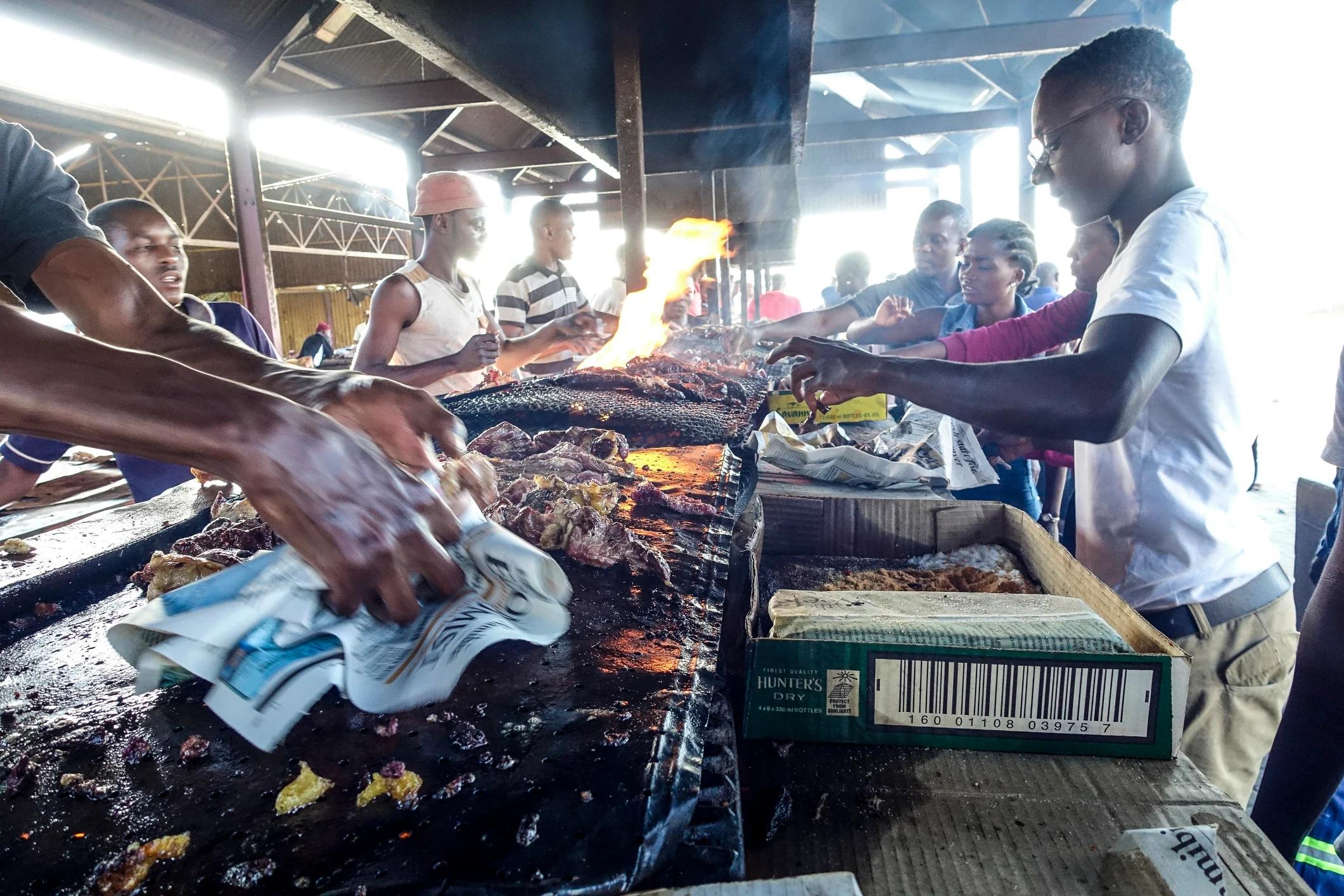 A bustling market scene with people grilling food over open flames, surrounded by vendors and onlookers.