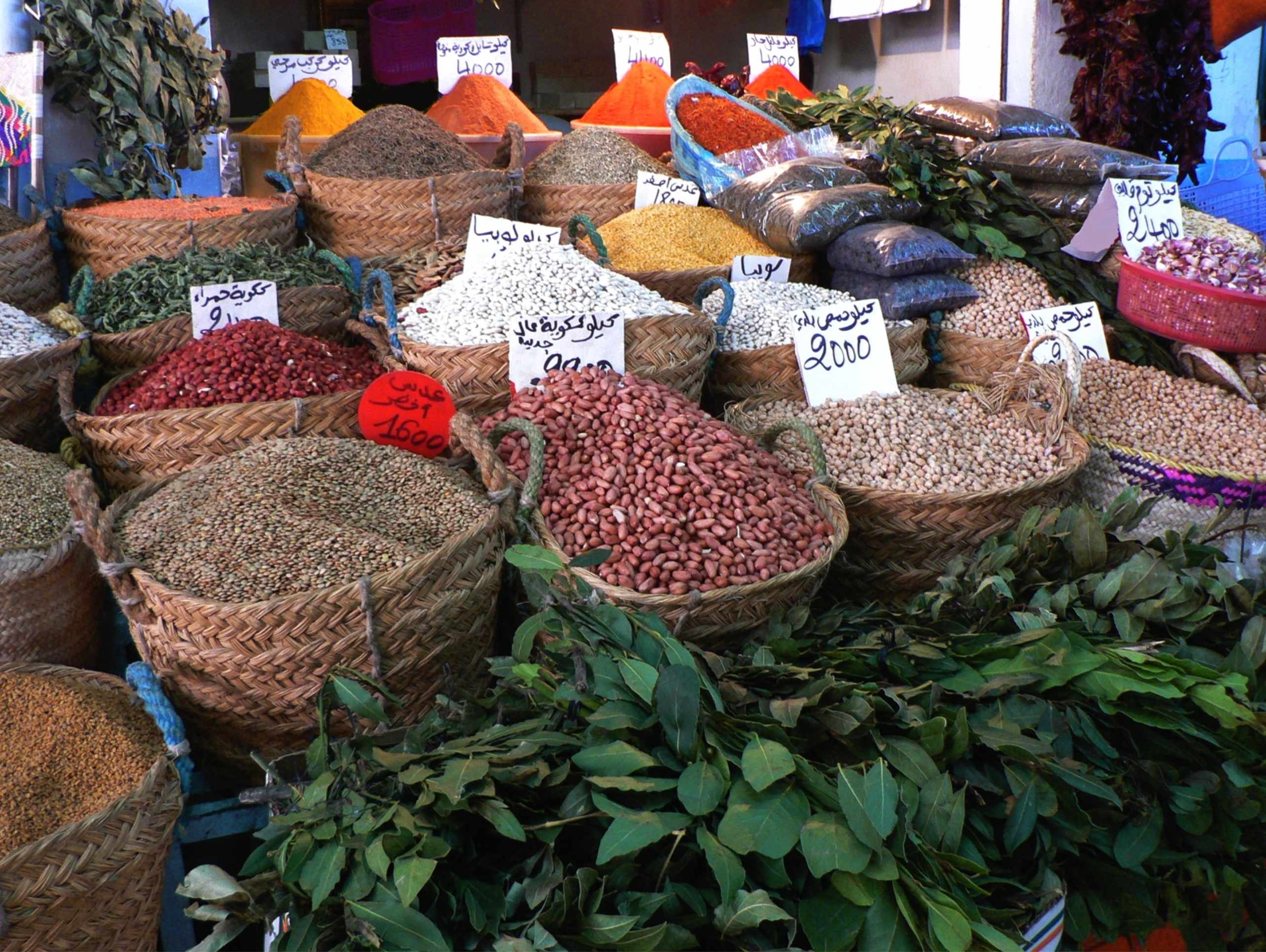 Baskets of colorful spices and legumes arranged in a market, with price tags visible in Arabic.