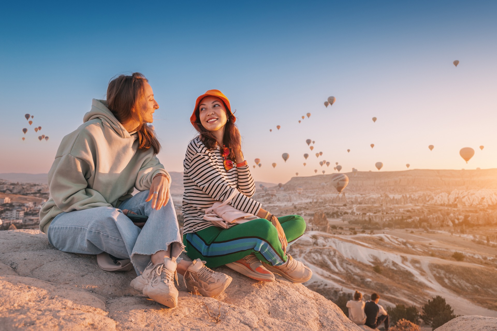 Two women sit on a cliff overlooking hot air balloons in Cappadocia, Turkiye.