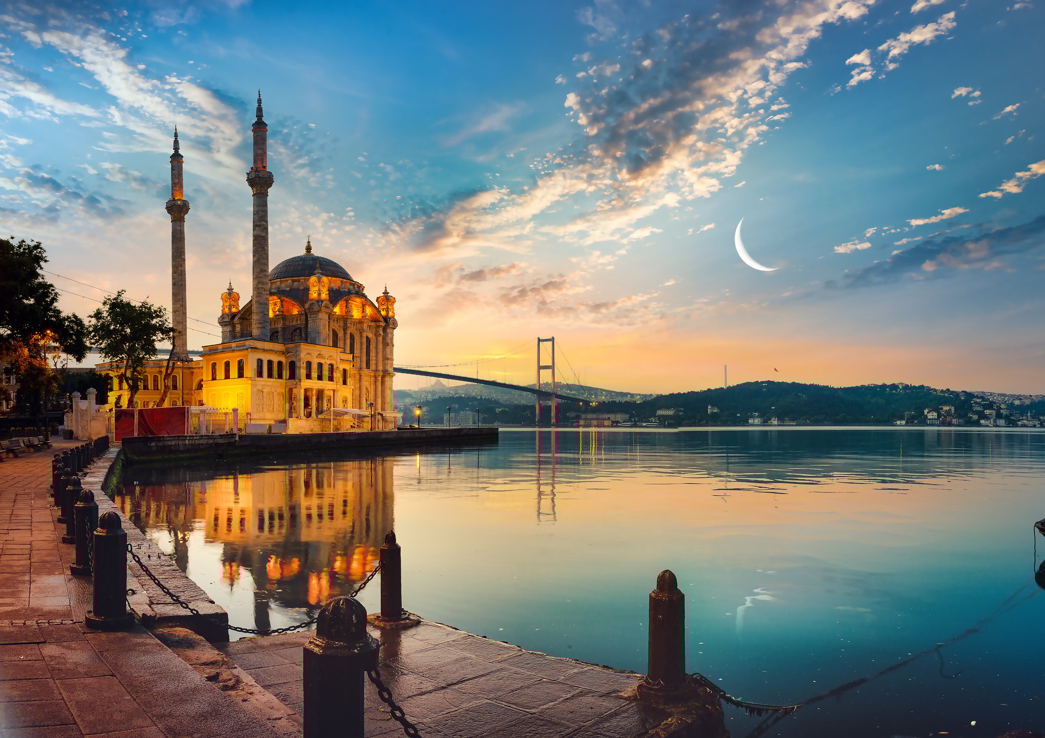 View of Ortakay Mosque in Istanbul with the Bosporus Bridge and the moon in the sky.