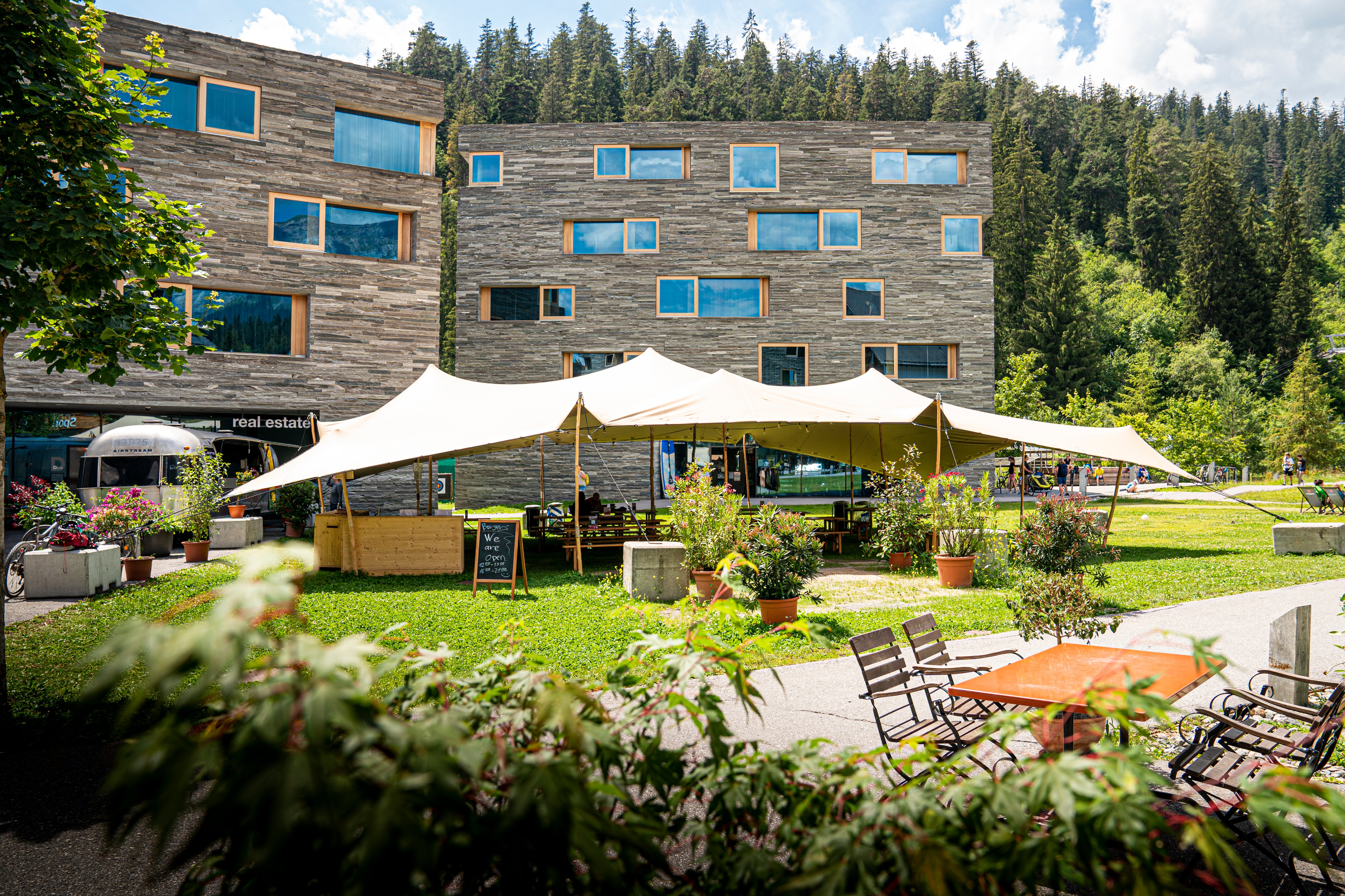 People relaxing in the shade beneath an open-air canopy tent, surrounded by several pots of colourful flowers and ferns, framed by two tall grey wooden cubic buildings and tall spruce trees in the background, in Laax, Switzerland.