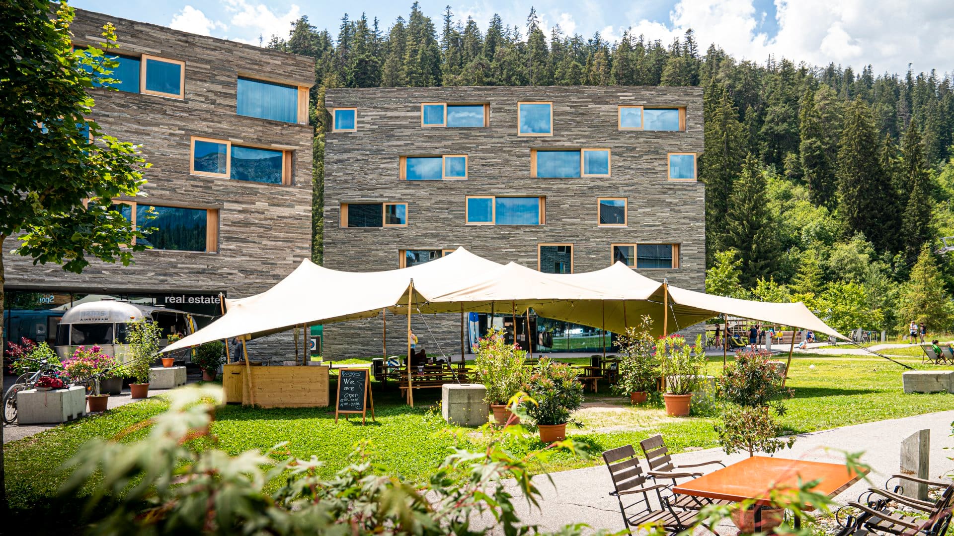 People relaxing in the shade beneath an open-air canopy tent, surrounded by several pots of colourful flowers and ferns, framed by two tall grey wooden cubic buildings and tall spruce trees in the background, in Laax, Switzerland.