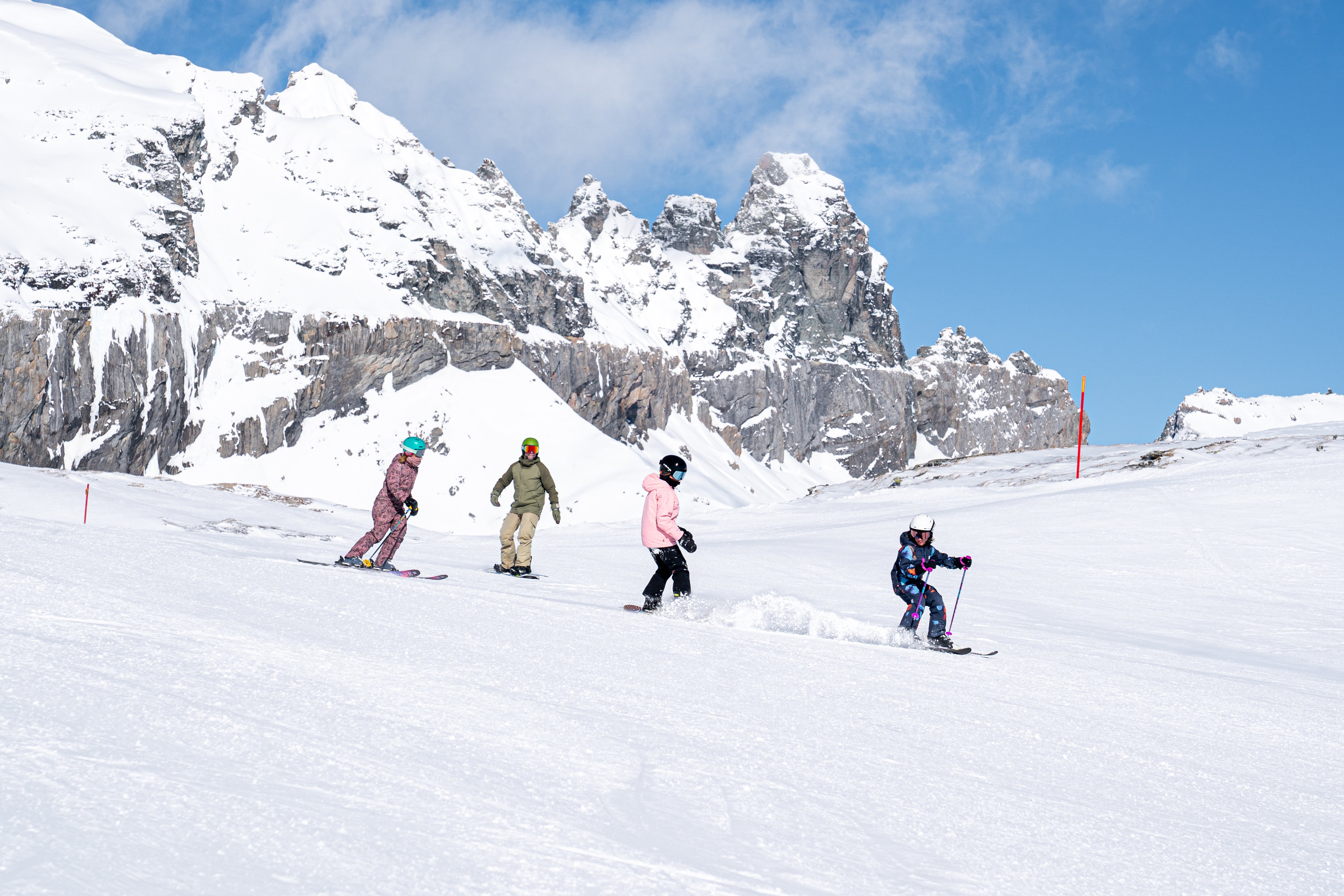 A family of four, with a child leading the way, skiing down a ski slope with ragged mountains behing them, in Laax, Switzerland.