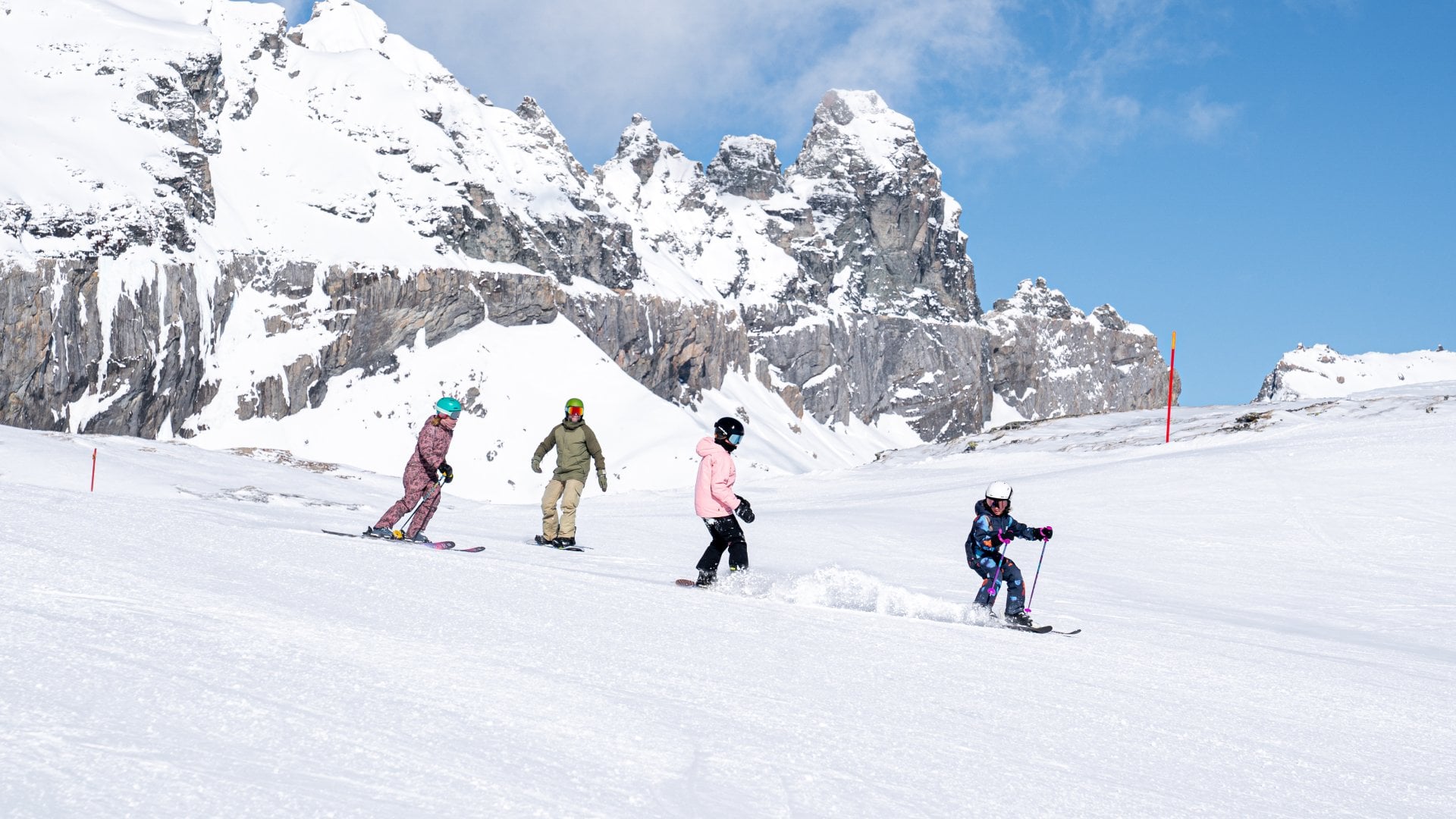 A family of four, with a child leading the way, skiing down a ski slope with ragged mountains behing them, in Laax, Switzerland.