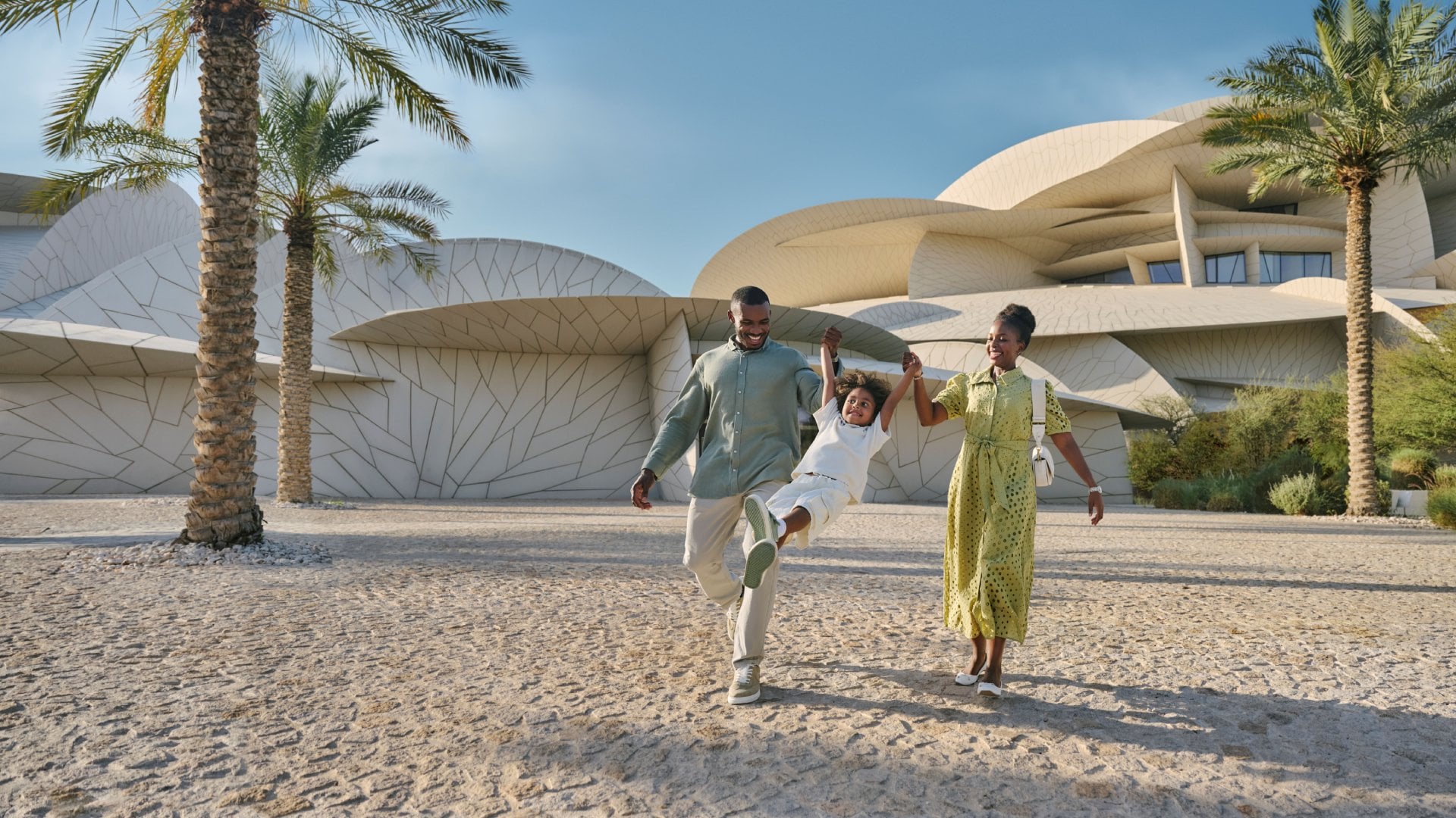 A mom and dad swing their child on the sands in front of an interesting building in Qatar