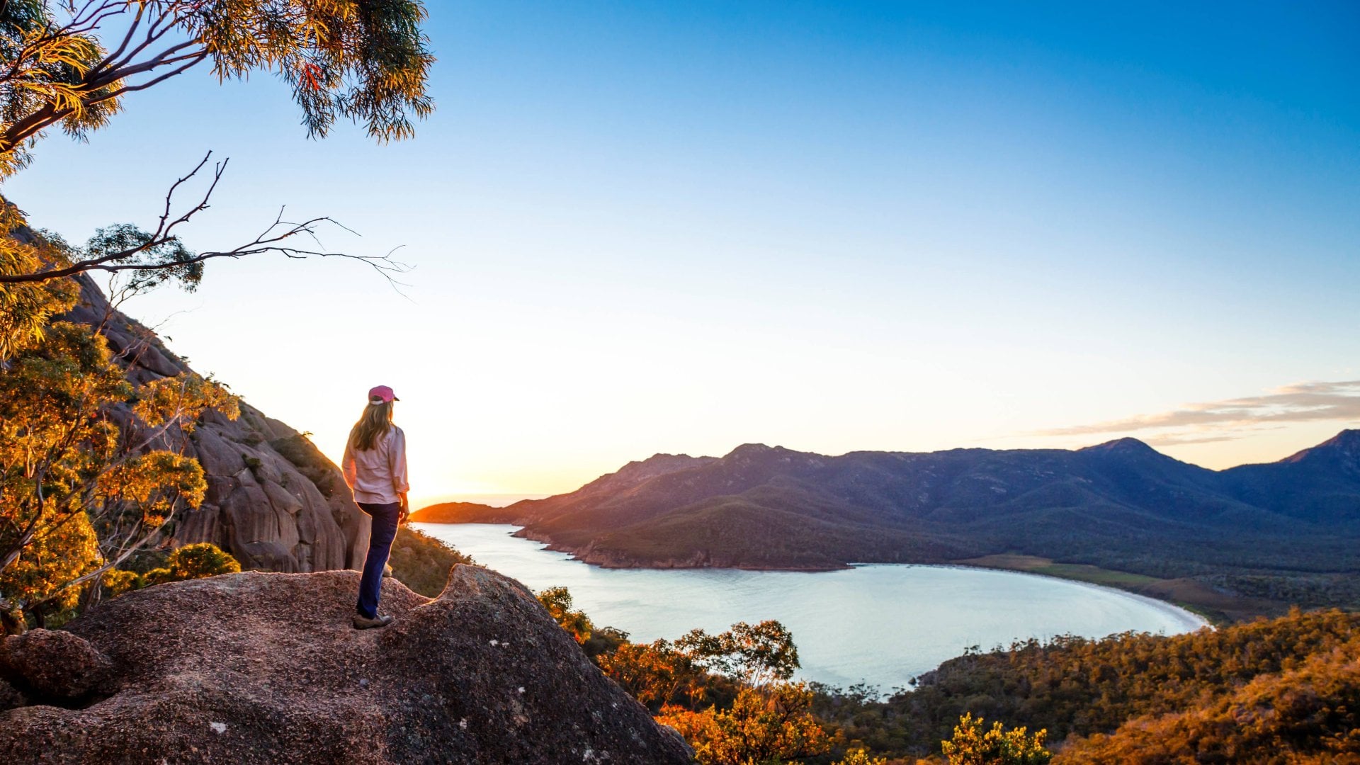 A woman standing on a rocky lookout point, watching the sun rise over Wineglass Bay in Freycinet National Park, Tasmania, Australia.