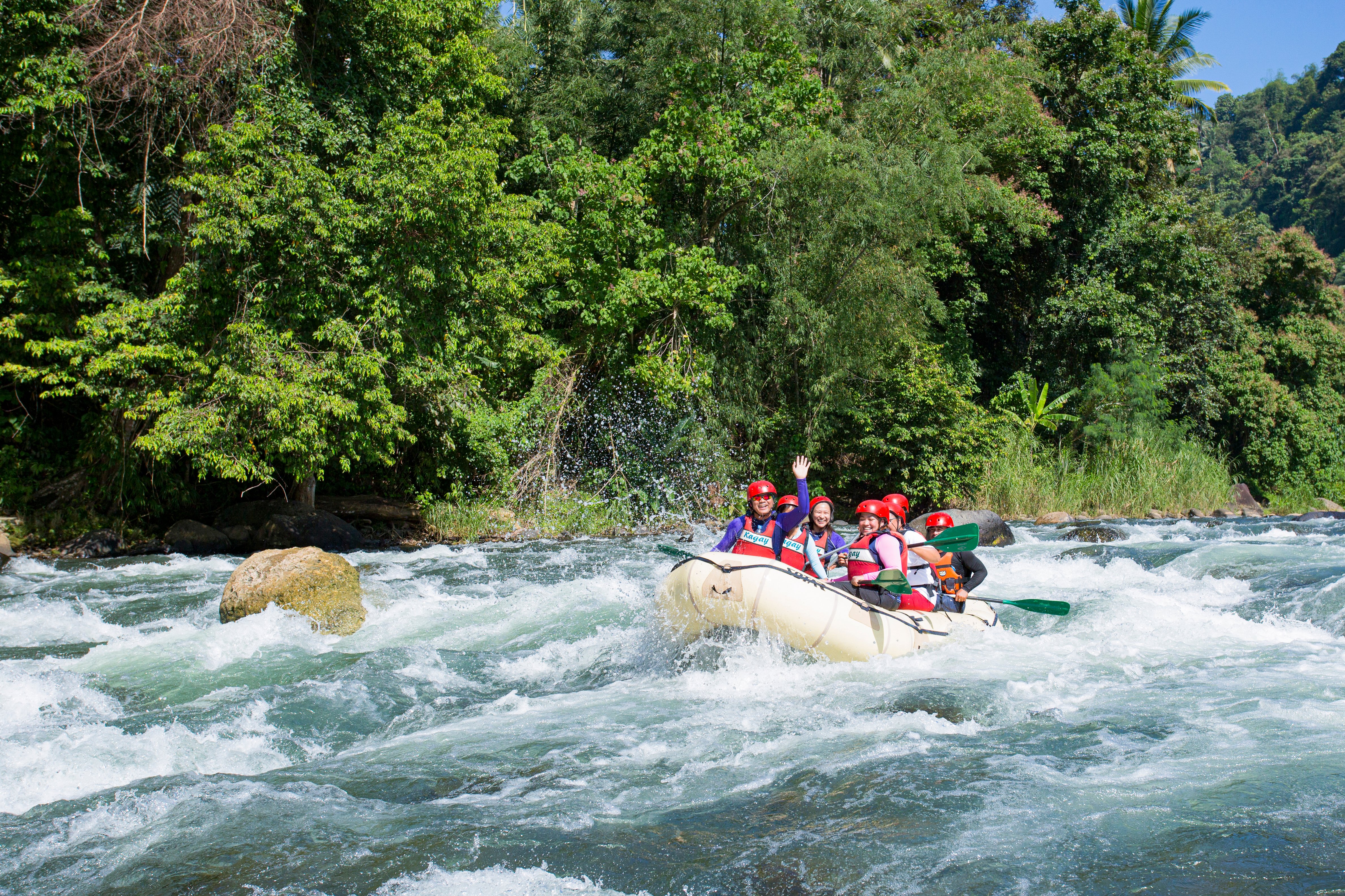 A group of people wearing red helmets and lifejackets, smiling and waving in a raft as they paddle down a river, with lush green trees and vegetation behind them in CAGAYAN DE ORO, Philippines.