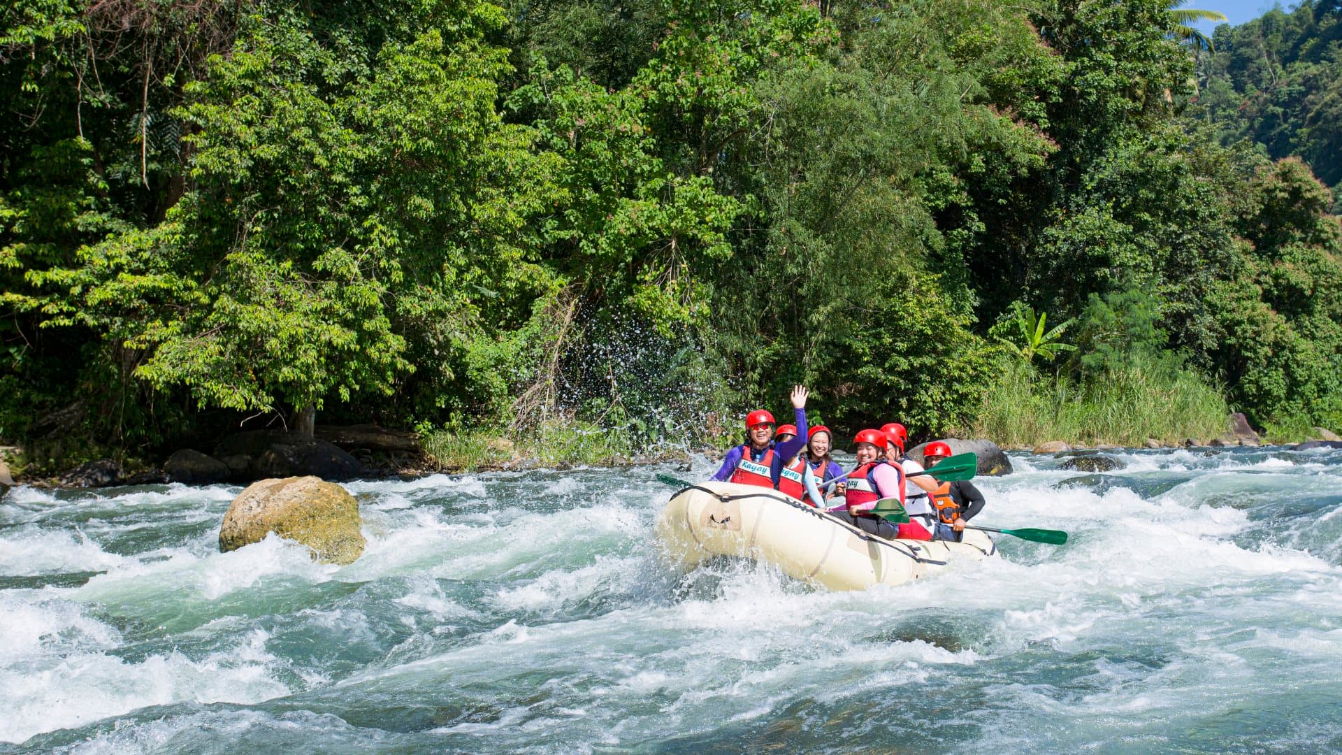 A group of people wearing red helmets and lifejackets, smiling and waving in a raft as they paddle down a river, with lush green trees and vegetation behind them in CAGAYAN DE ORO, Philippines.