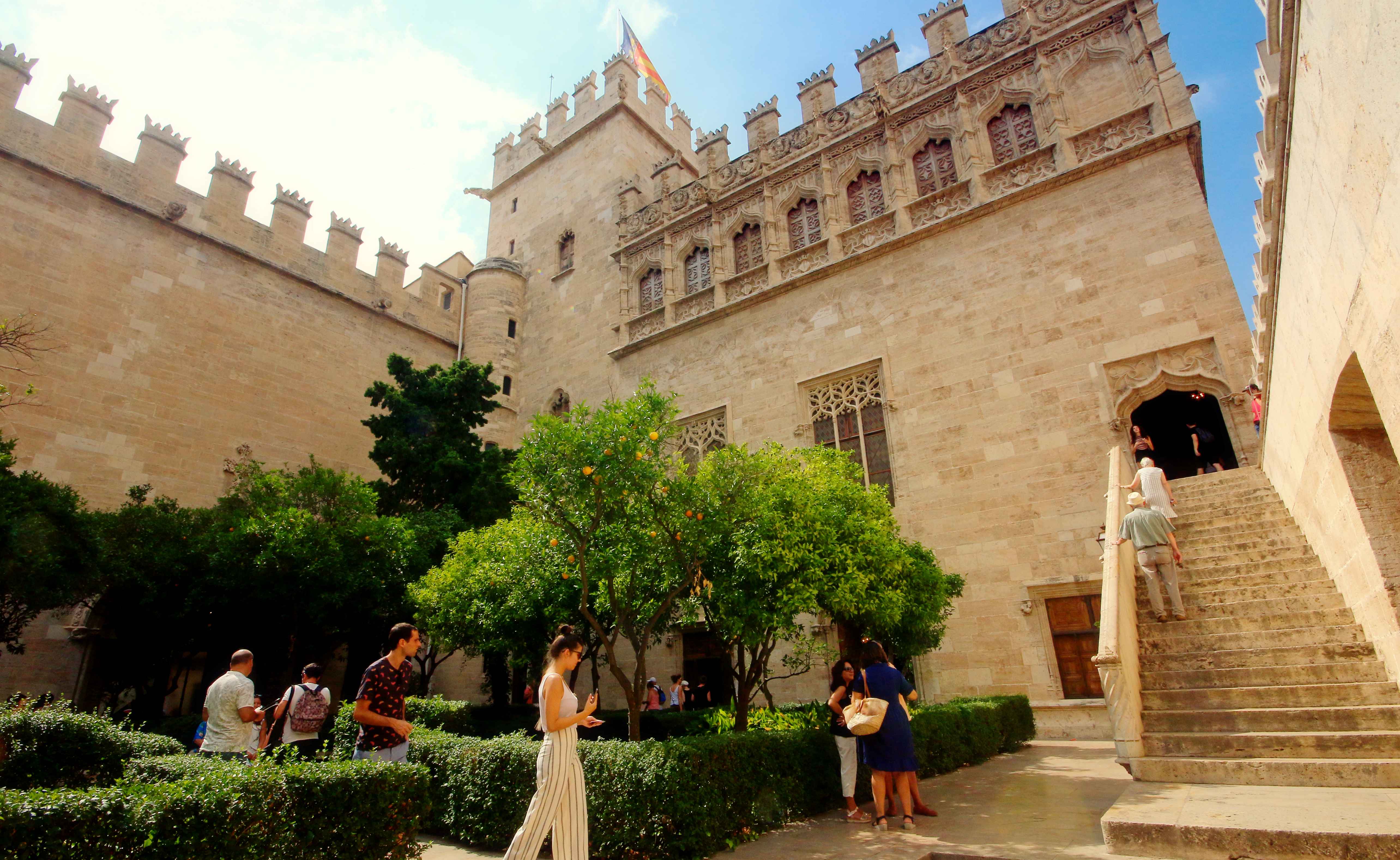 A group of travellers in the bottom-left frame walk in a green, leafy area of an outdoor courtyard surrounded by the towering crenellated walls of the Silk Exchange UNESCO World Heritage Site in Valencia, Spain.
