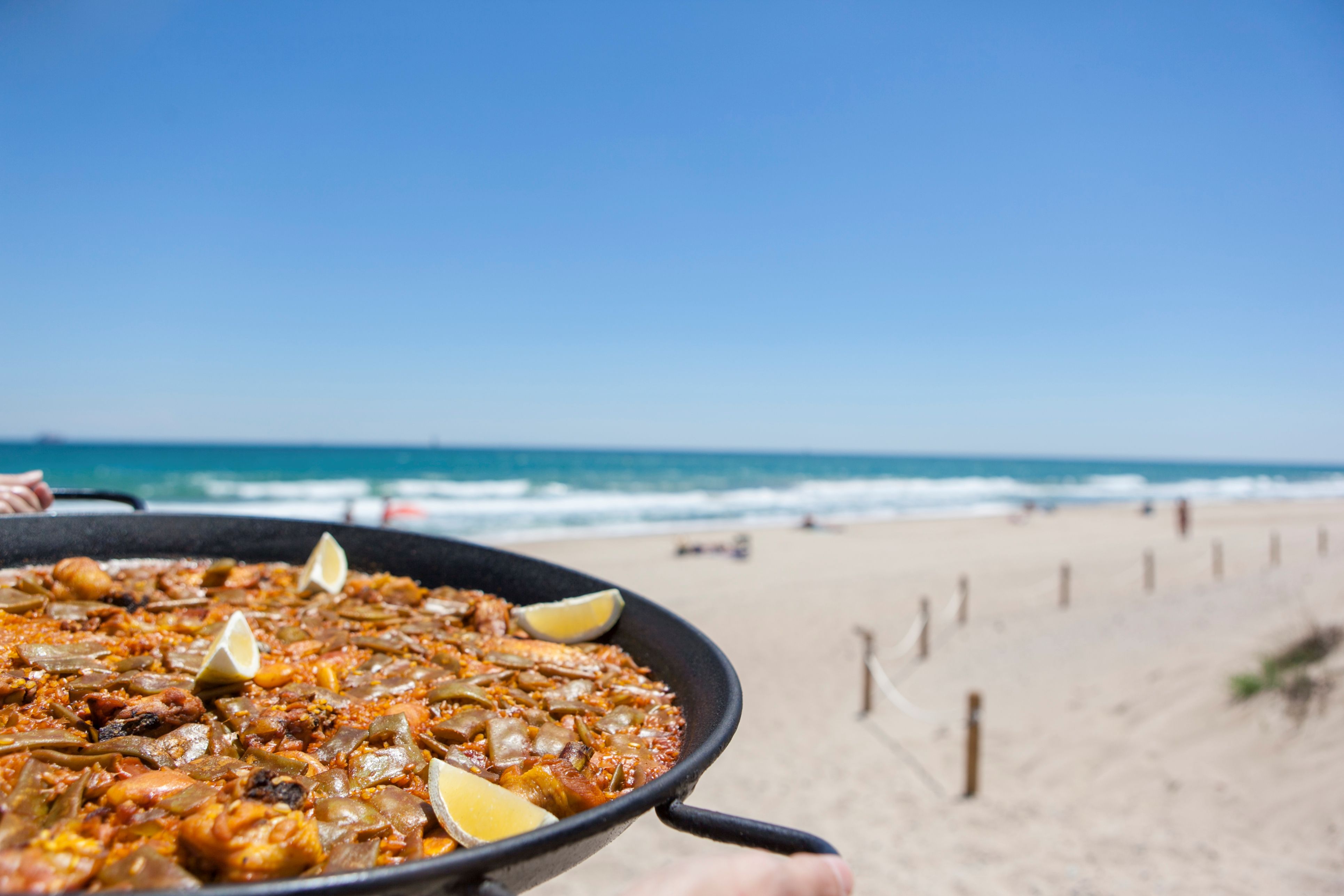 A scrumptious dish of traditional Valencian paella covers the bottom-left frame, with a sunny white-sand beach with a bright blue ocean and sky in the background.