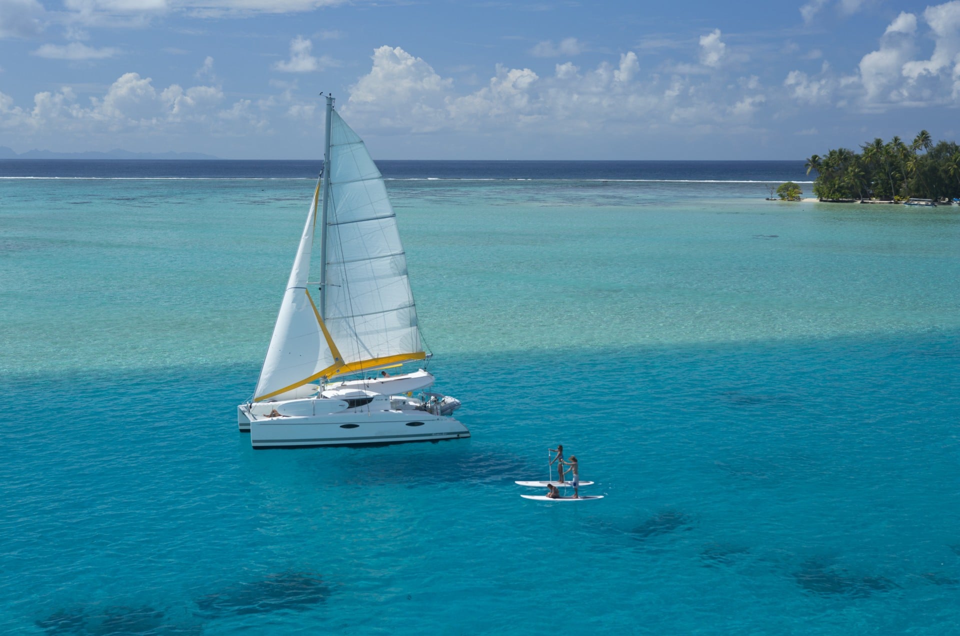 Two paddleboarders and a catamaran in a blue lagoon in Tahiti.