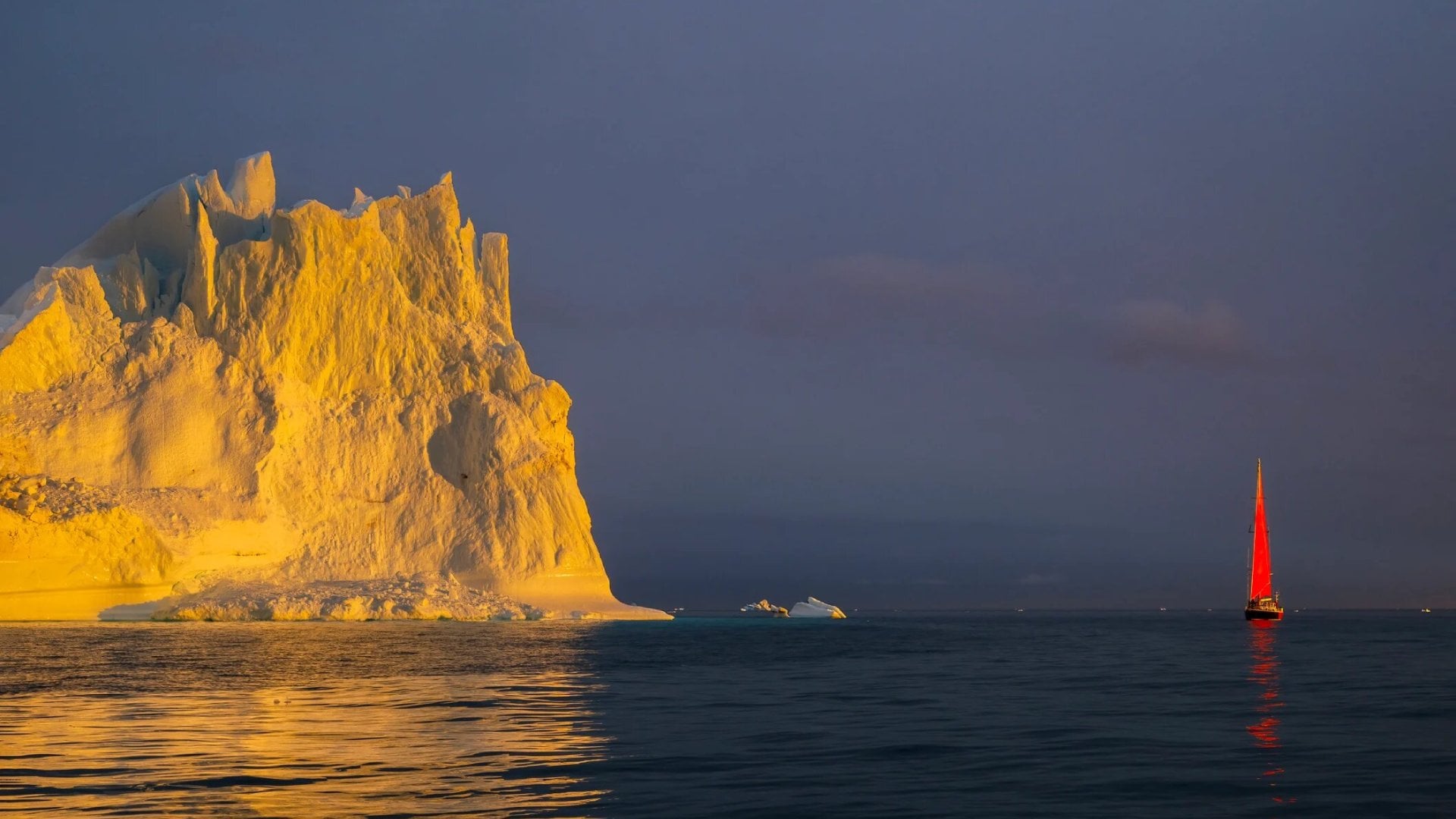 Red sailboat sailing among floating yellow icebergs hit by setting sun in Disko Bay during polar summer midnight solar season. Ilulissat, Greenland