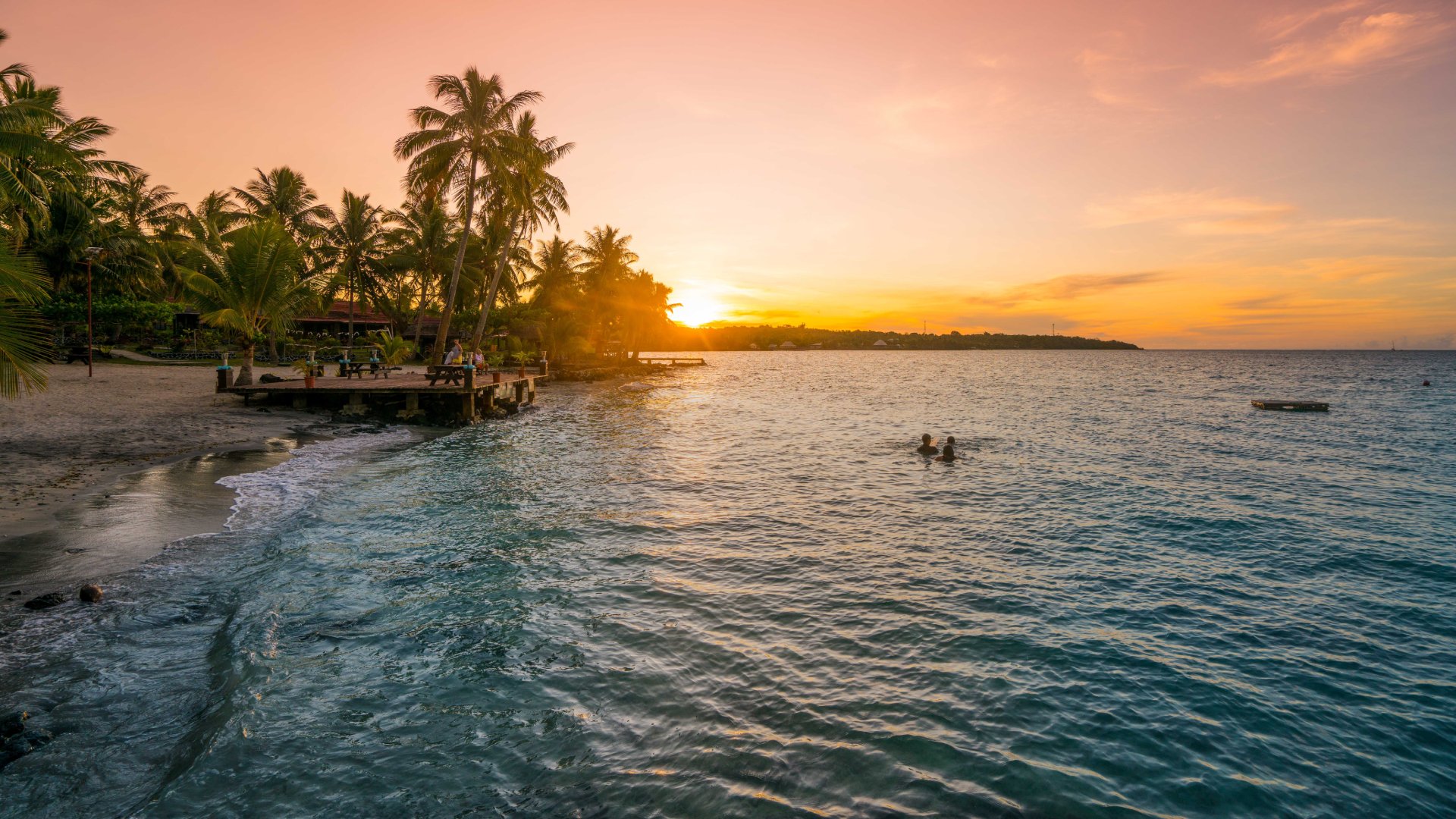 A group of people watching two other people swim off a beach at sunset.