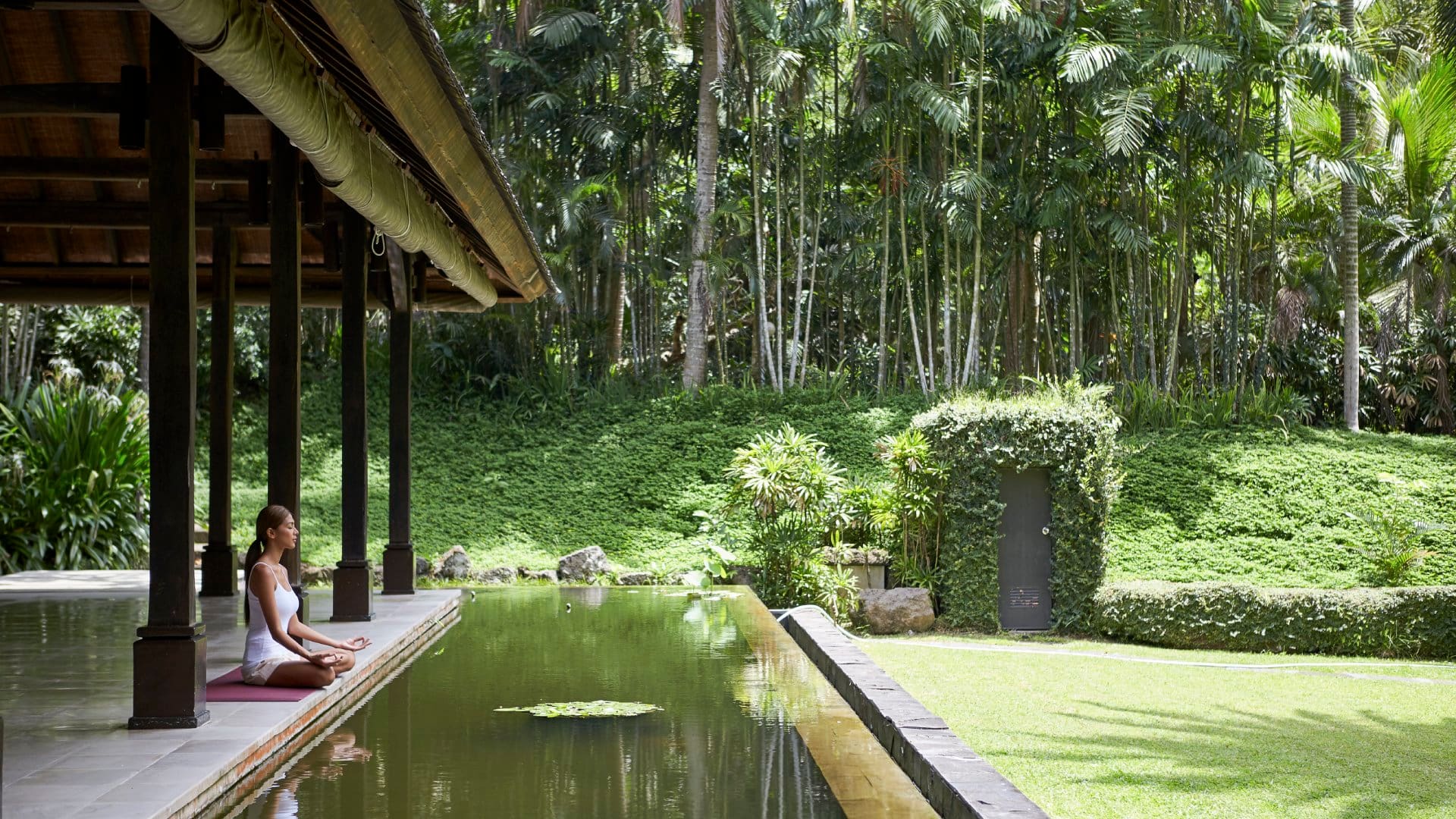 A woman on the left side of the image is sitting down in a cross-legged yoga pose, meditating beneath an open-air roof in a lush jungle setting in the Philippines.
