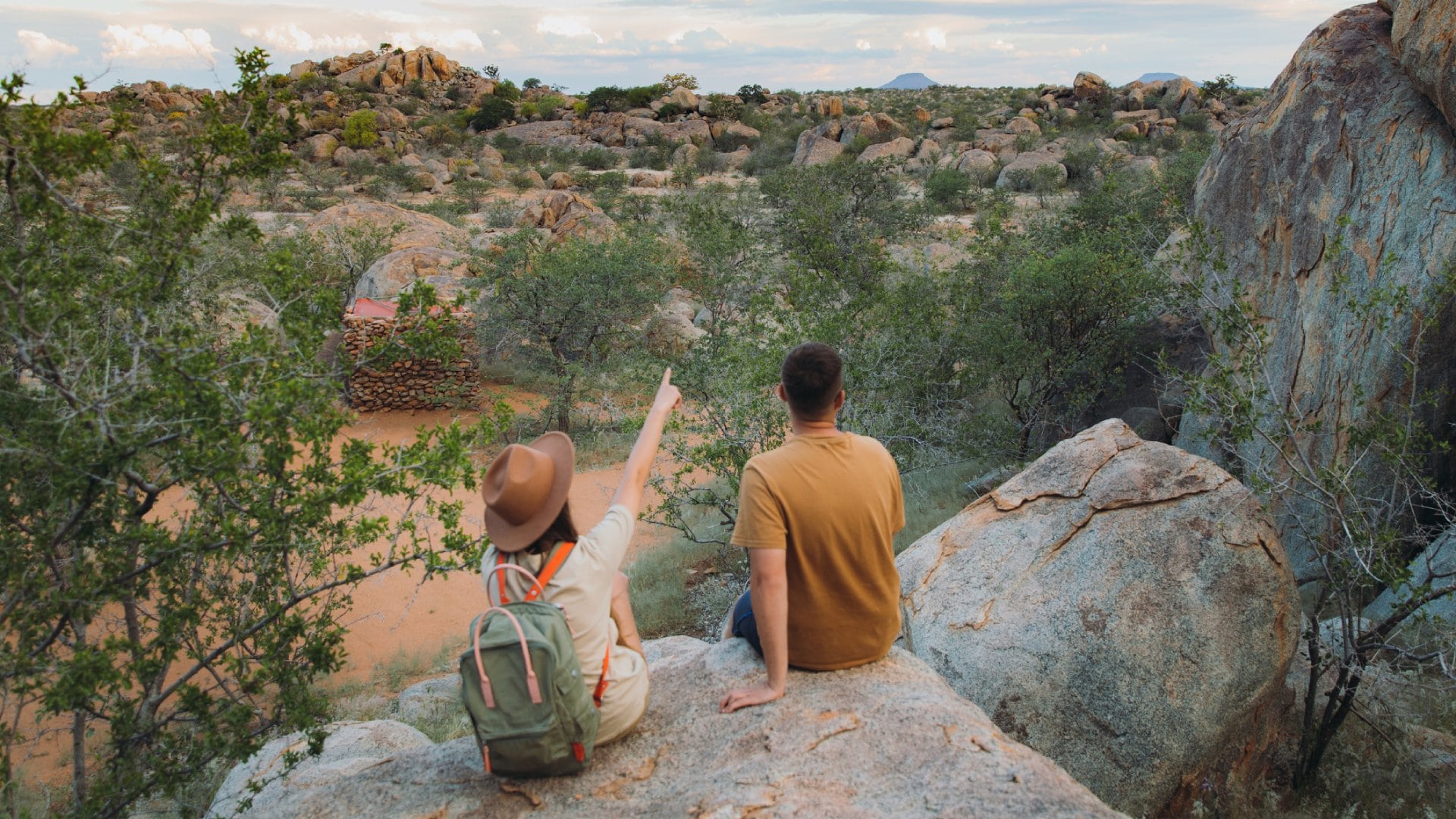 Young couple of woman in hat with backpack and man sitting on the top of the rock looking at the beautiful landscape and watching wild animals in Damaraland, Namibia, Southern Africa