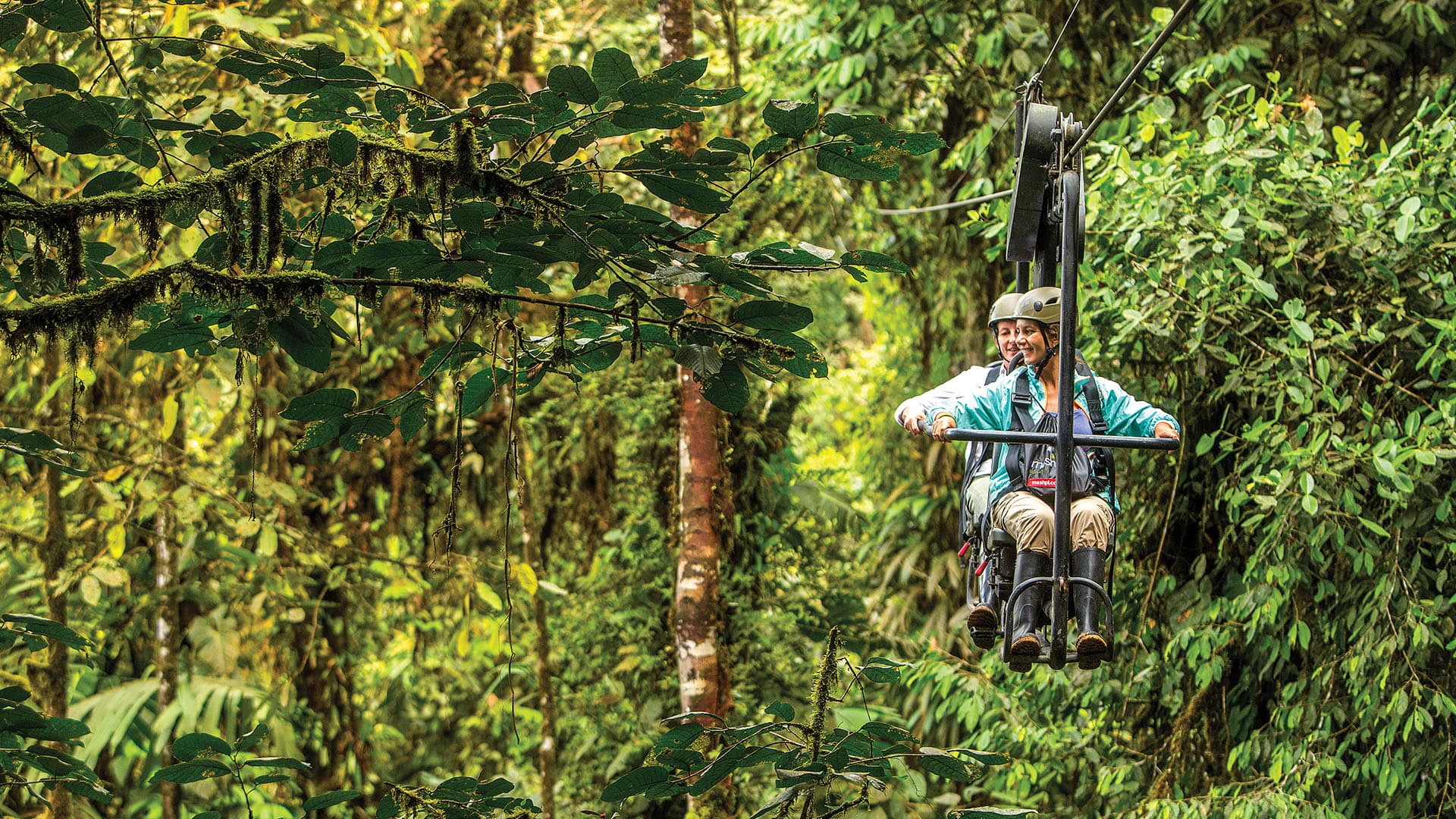 Two people seated on a zipline going through a lush green jungle canopy