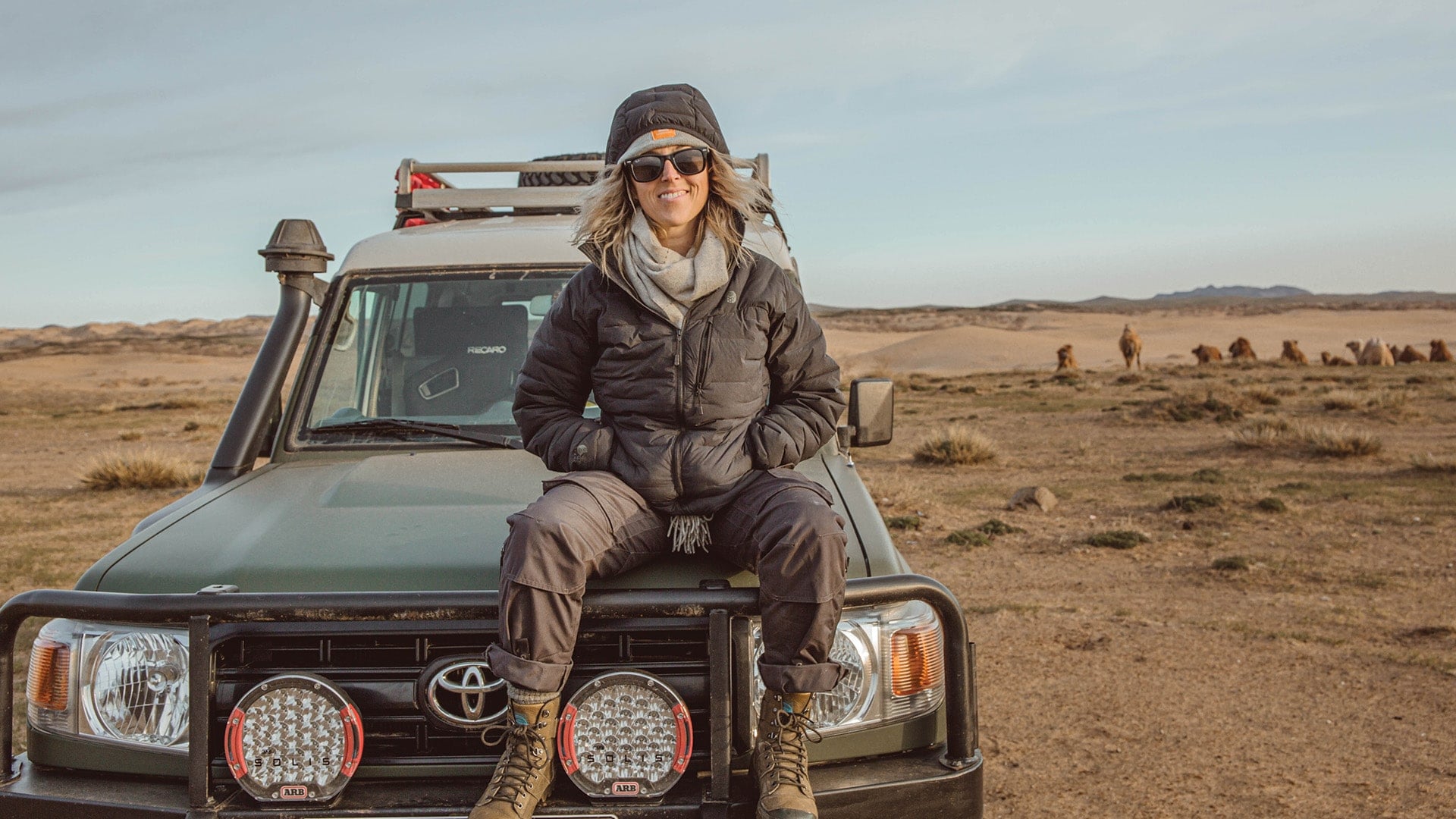 Breanna Wilson sitting on the hood of her custom-built Landy Cruiser 78 Troopy next to a group of camels in the desert