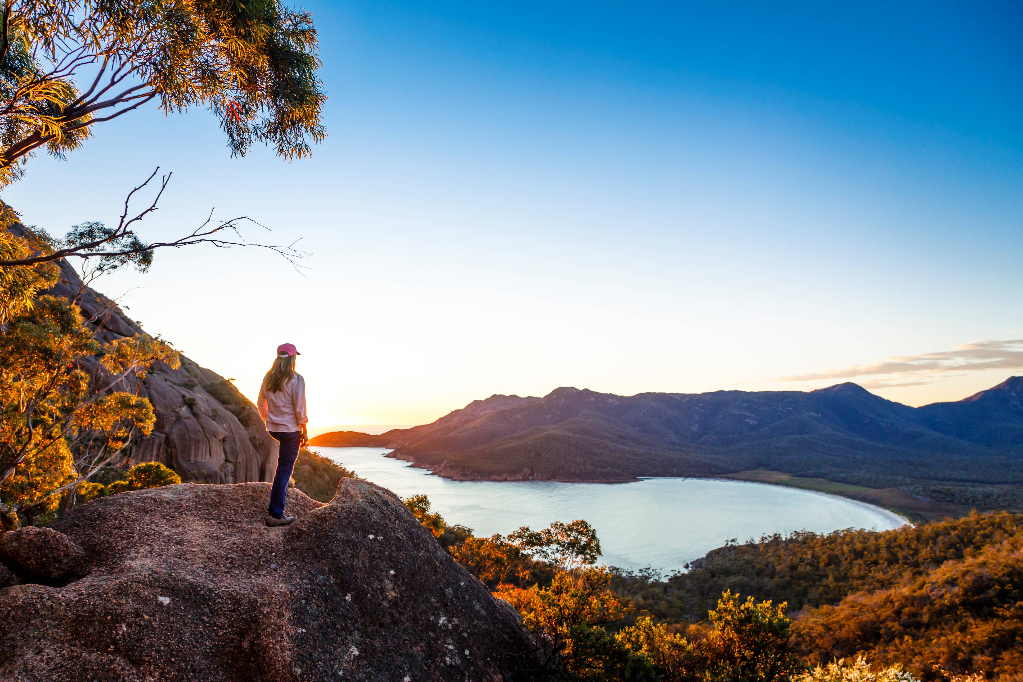 A woman standing on a rocky lookout point, watching the sun rise over Wineglass Bay in Freycinet National Park, Tasmania, Australia.