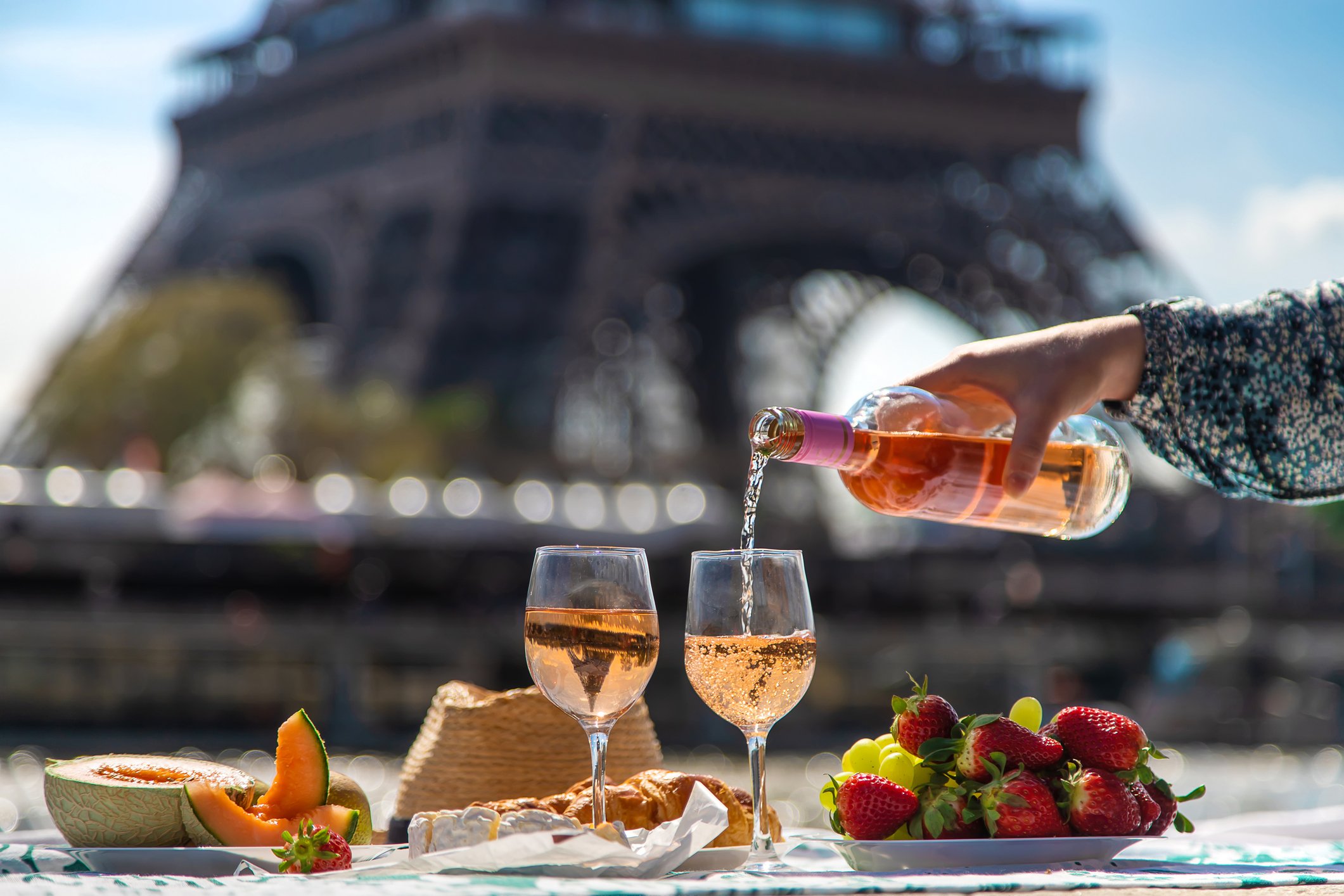 A woman sitting off-screen pouring rose into two wine galsses, with a bowl of strawberries and grapes, a canteloupe, a croissant, and cheese to the side, with the Eiffel Tower in the background in Paris, France