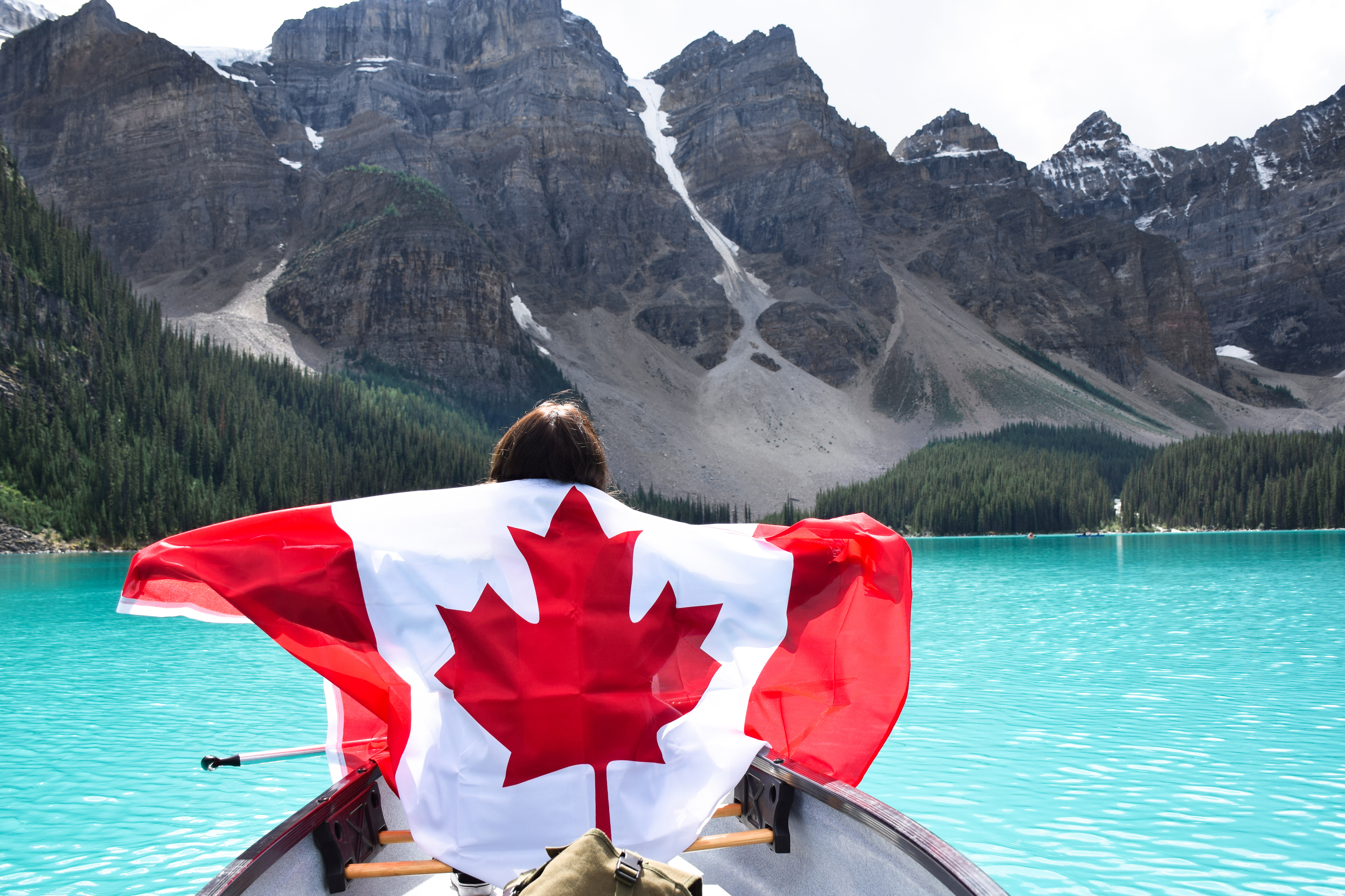 A woman holding a Canadian flag across her back as she canoes through the bright turquoise waters of Lake Moraine, Canada.