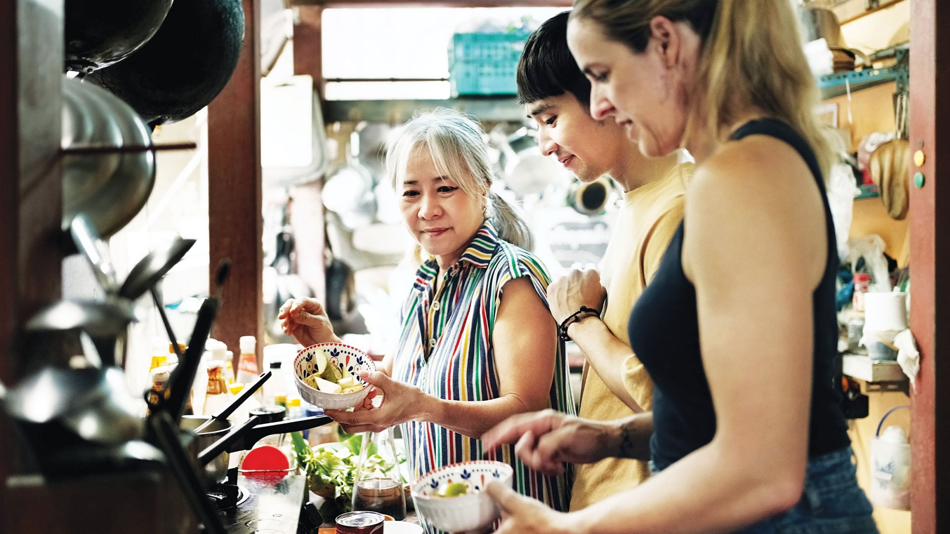 Two tourists take part in a cooking class with an older woman.