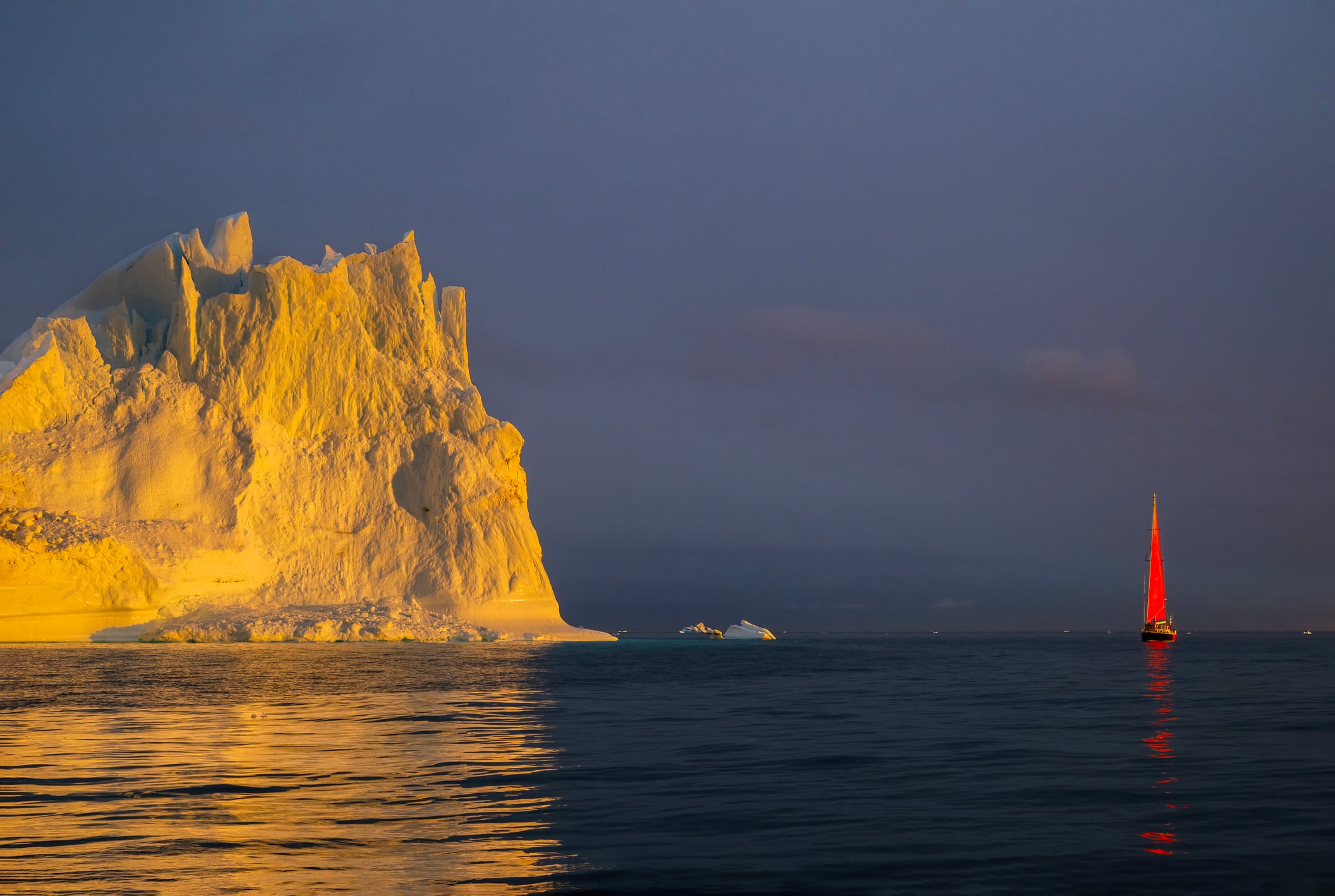 Red sailboat sailing among floating yellow icebergs hit by setting sun in Disko Bay during polar summer midnight solar season. Ilulissat, Greenland