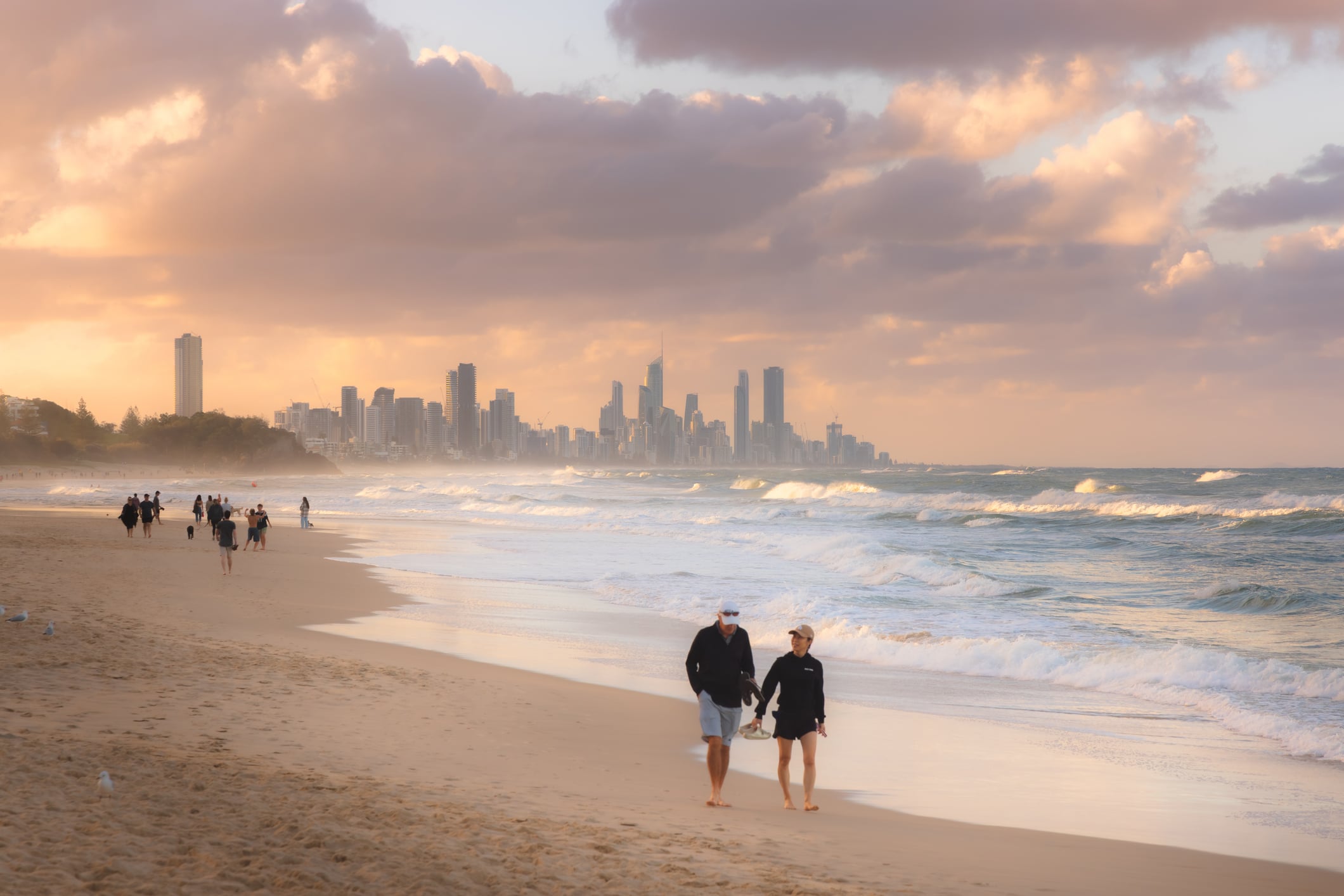 A couple walking along a beach in Burleigh Heights, Gold Coast, Australia, with other people in the background and an orange, cloudy, sunset sky above.