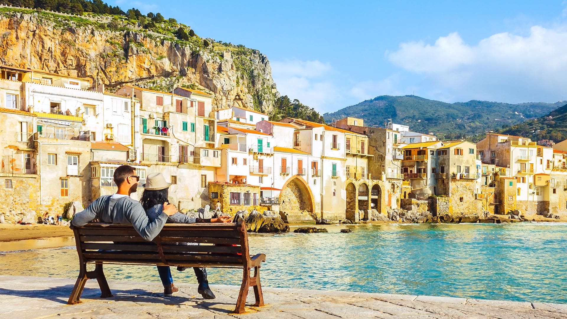 a couple relaxes on a bench gazing out over the golden homes lining the waters on an idyllic day in Sicily